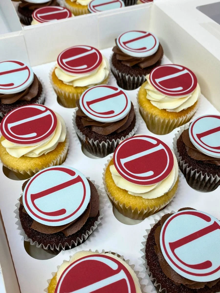 Assorted cupcakes with white, yellow, and chocolate frosting, topped with edible images of the number 10 in red and white, arranged in a white box.