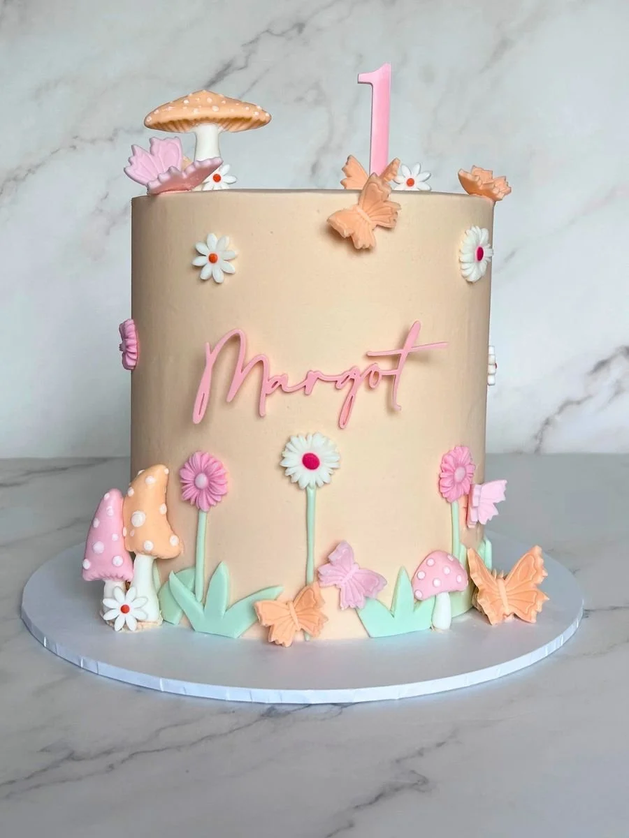 A pastel-colored birthday cake decorated with flowers, butterflies, and mushrooms. The cake has a pink 'Margot' inscription and a pink number one candle on top, set against a marble background.