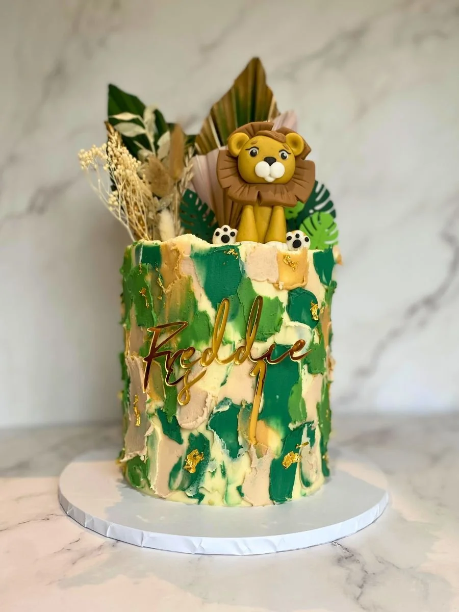 Decorative birthday cake with a jungle theme, featuring a lion topper, green marbleized frosting, gold accents, and dried foliage decorations. The cake has the name 'Freddie' written on it.