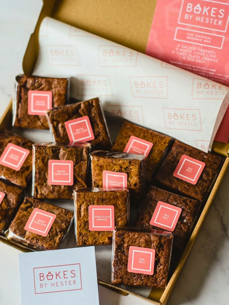 Assorted baked brownie squares with pink labels inside a box, from Bakes by Hester, featuring flavors such as chocolate orange, white choco chip, salted caramel, chocolate orange, and sea salt.