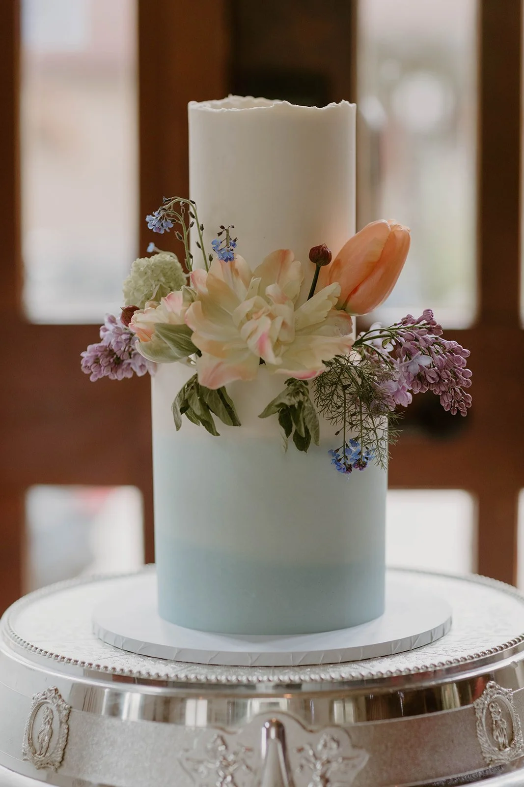 A white cylindrical cake decorated with a variety of pastel-colored flowers, including tulips and smaller purple and blue blossoms, placed on a decorative silver stand.