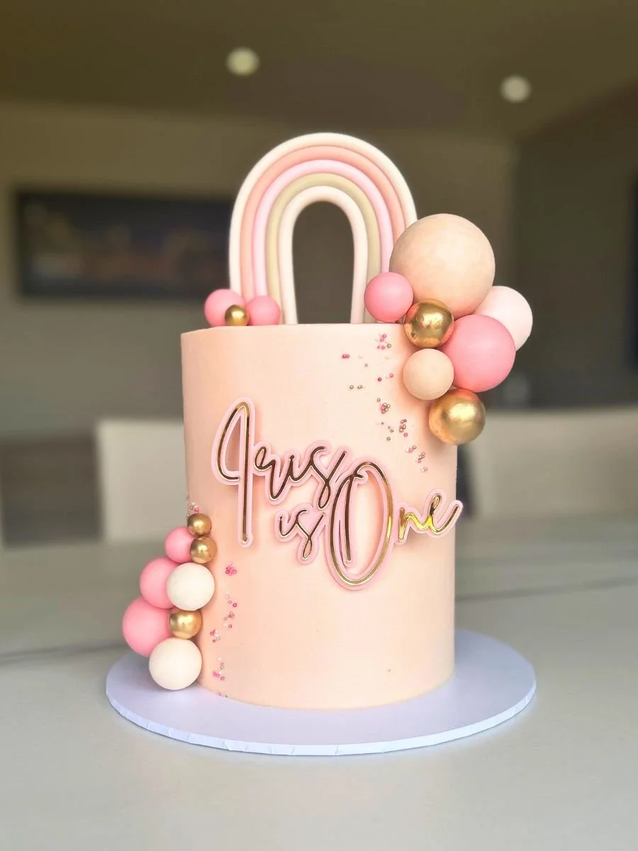 Pink cake shaped like a lock with a rainbow decoration on top, decorated with pink, white, and gold balls, and a "Trust & One" sign on the front.