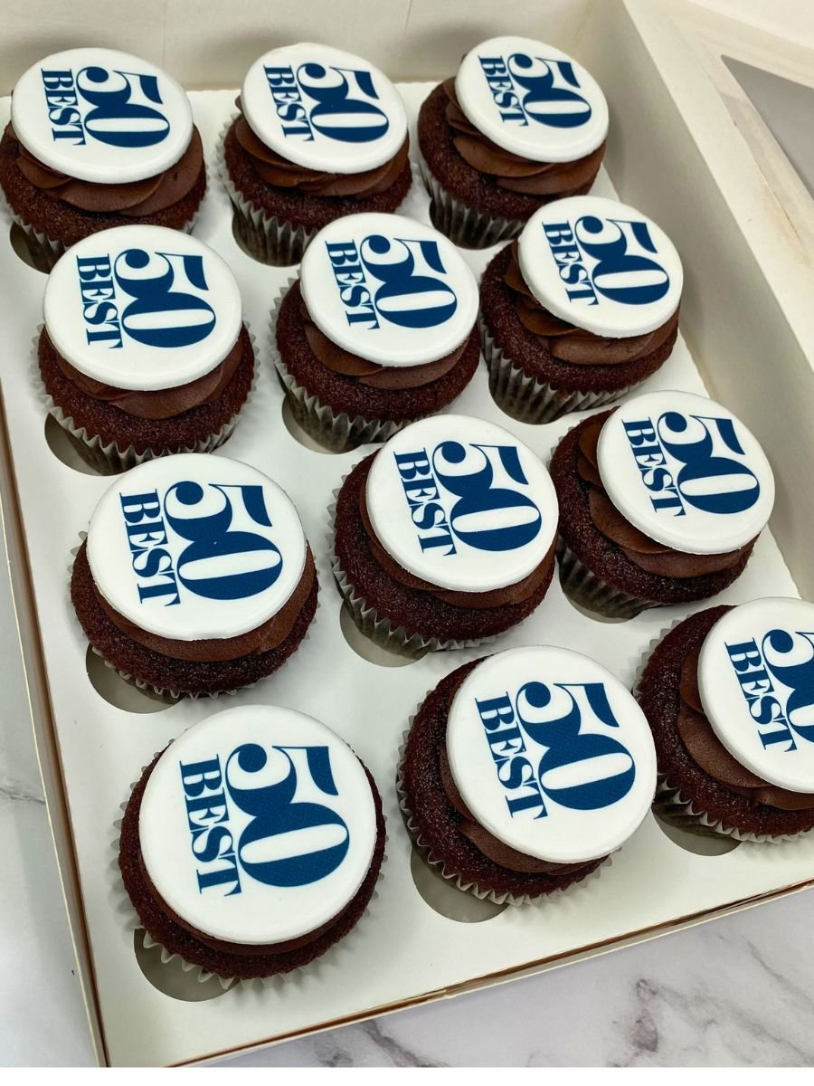 A box of cupcakes with white fondant toppers that say '50th Best' in blue text, celebrating a 50th anniversary or milestone.