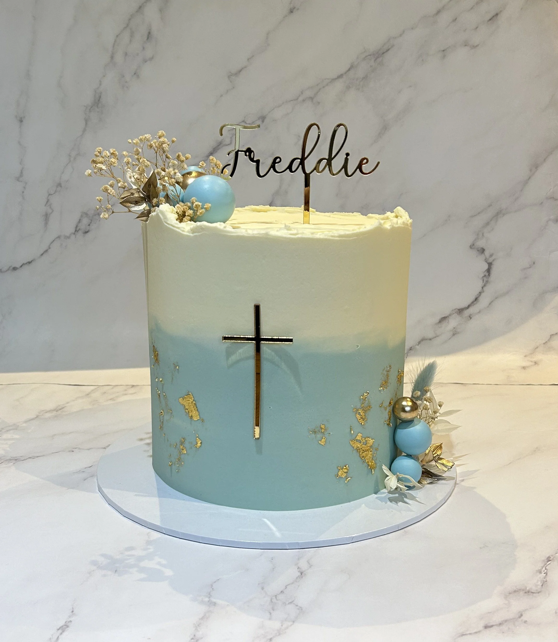 A cake with a gold cross on the front, decorated with white and light blue flowers, a blue ball, and a cake topper with the name 'Freddie'. The cake has a smooth white fondant finish with gold accents, set on a marble surface.