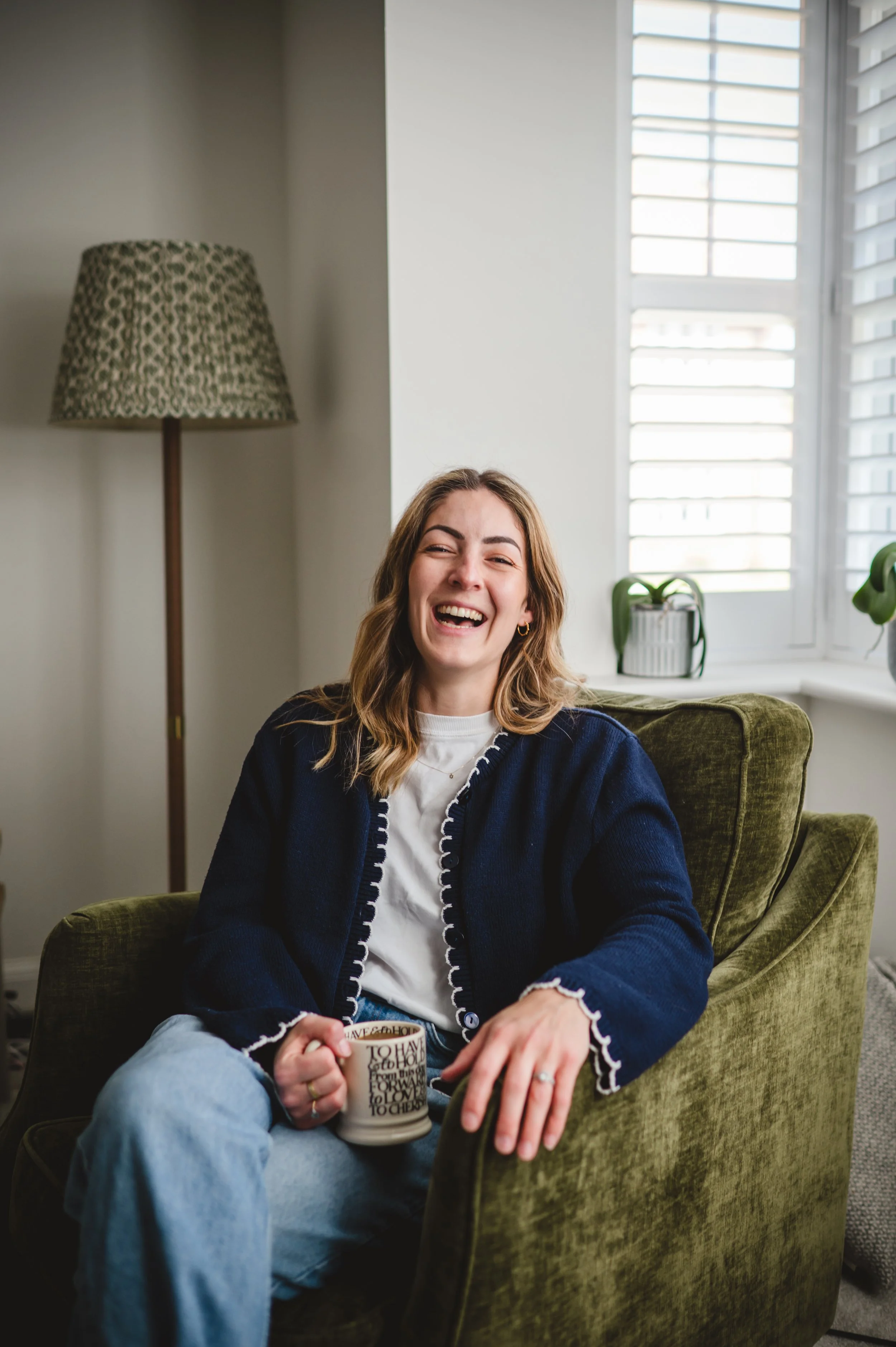 A woman sitting comfortably on a green armchair, smiling and laughing while holding a coffee mug with words on it, inside a bright room with white walls, a window with blinds, a potted plant, and a tall floor lamp with a patterned lampshade.