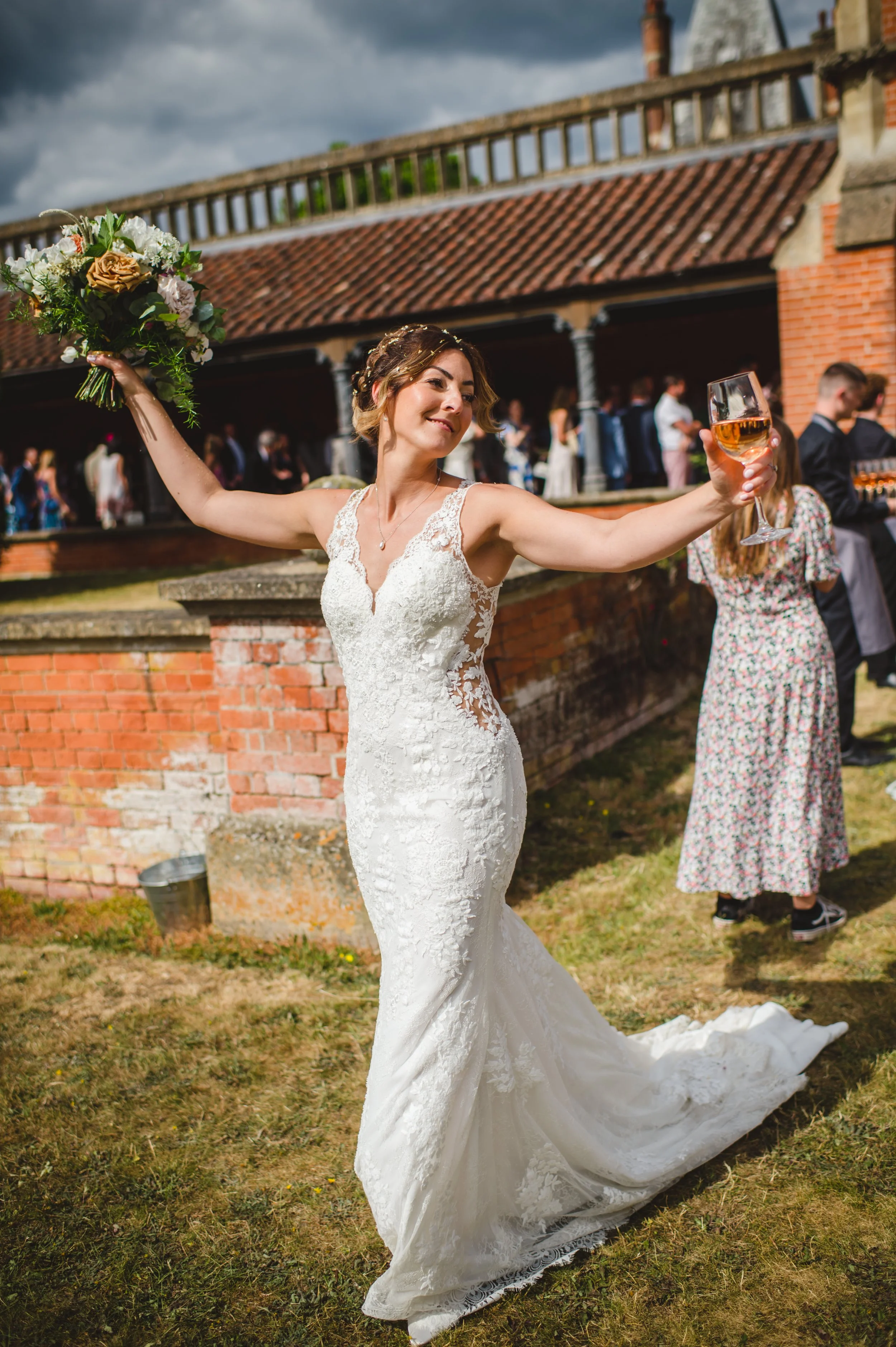 A bride in a white lace wedding dress holding a bouquet in her left hand and a glass of wine in her right, standing outdoors in front of a brick building with a red tile roof, with guests in the background.