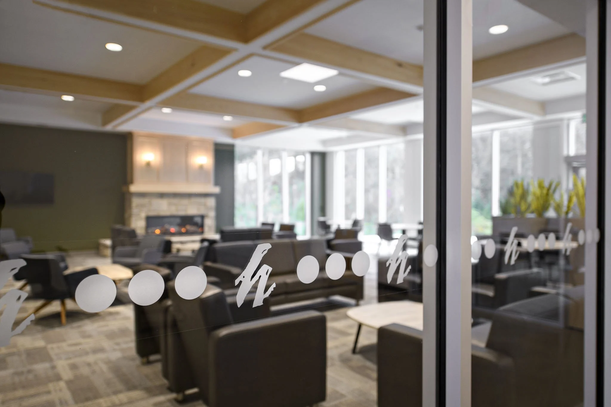 Modern lounge area with black armchairs, large windows, and a fireplace on a gray wall inside a building.