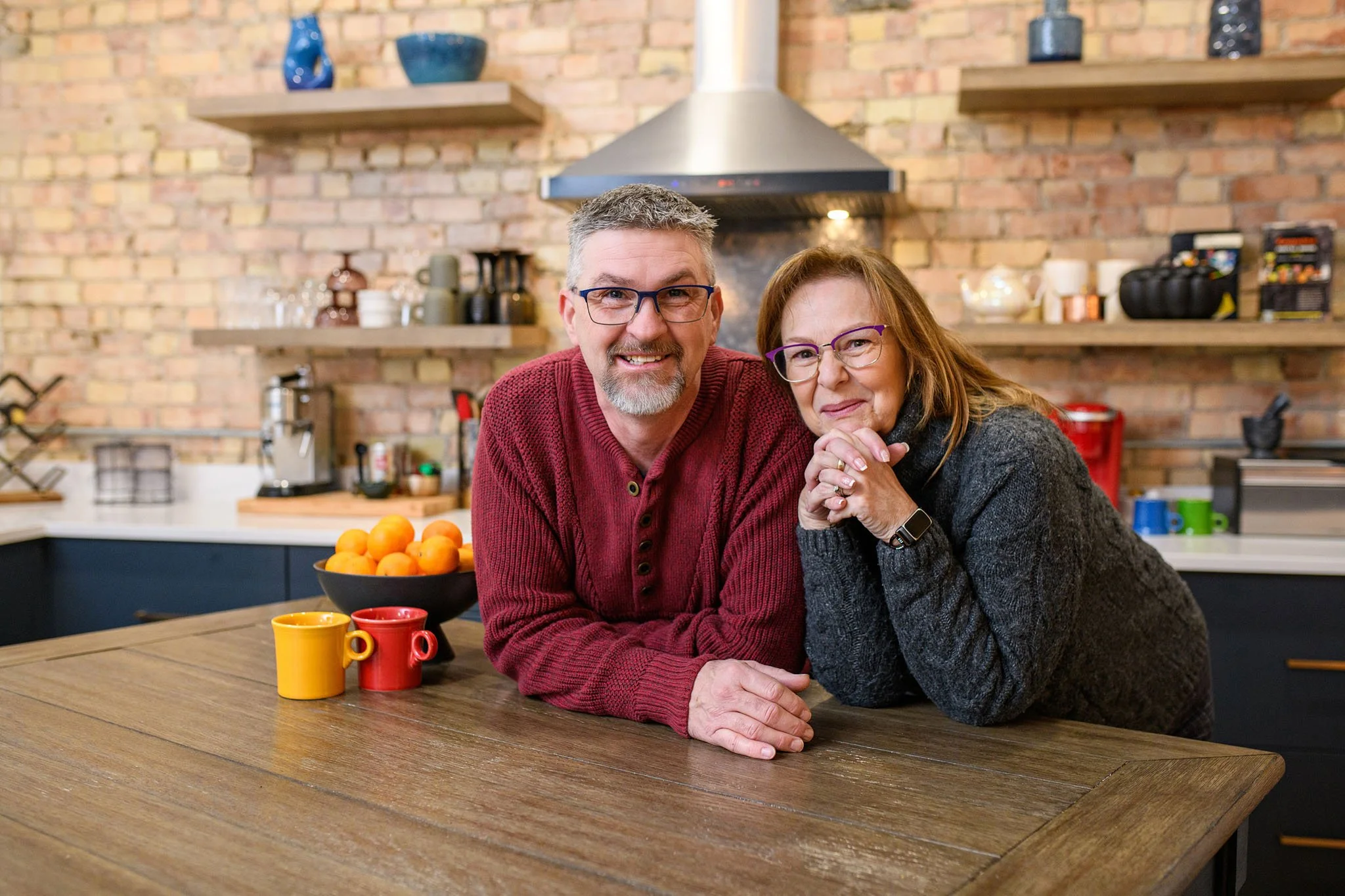 A happy middle-aged couple with glasses, smiling, leaning on a wooden kitchen table with orange, yellow, and red mugs, and a bowl of oranges, in a modern kitchen with brick walls and open shelves.