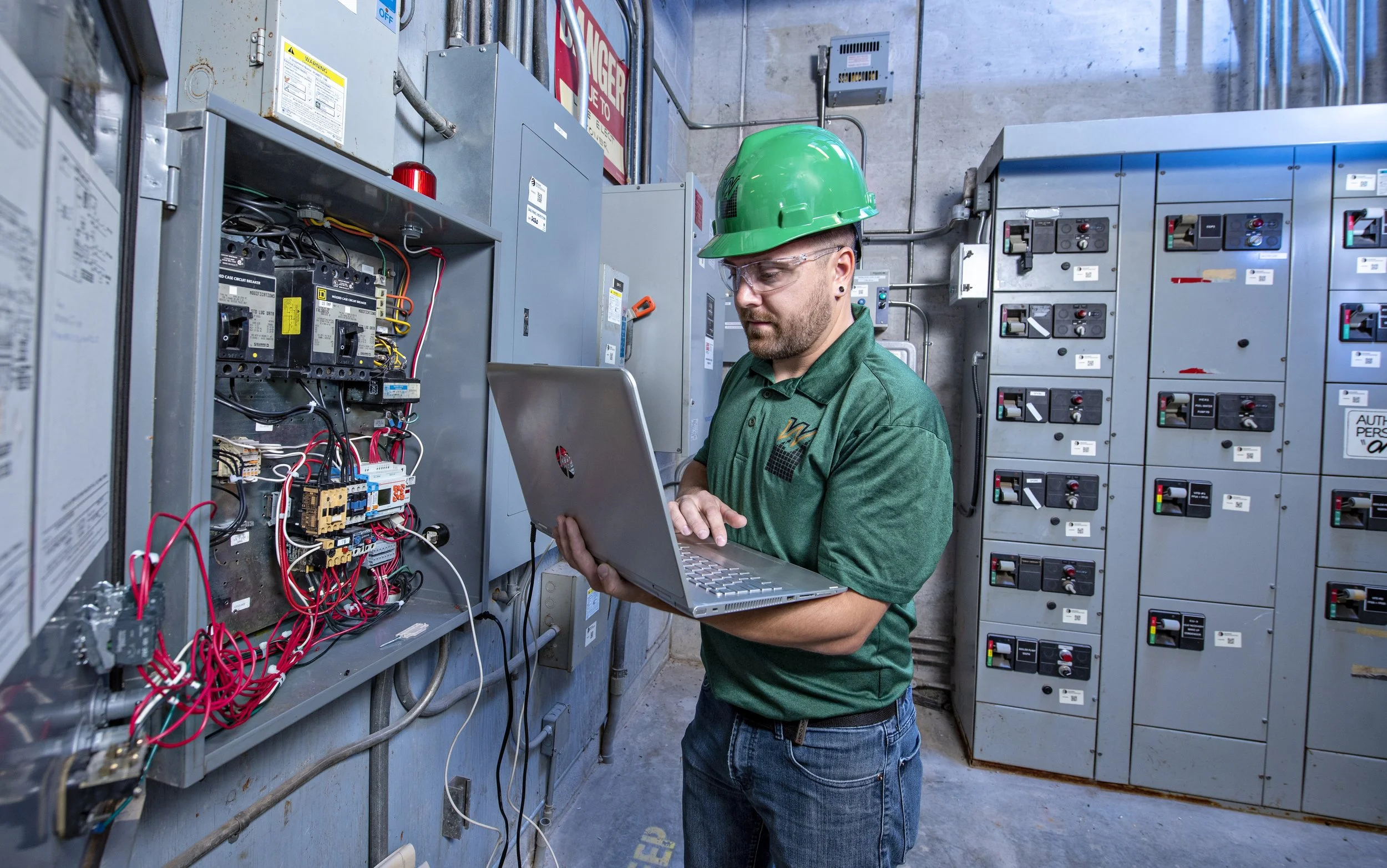 A technician wearing a green hard hat and safety glasses working on electrical panels with a laptop in an industrial setting.