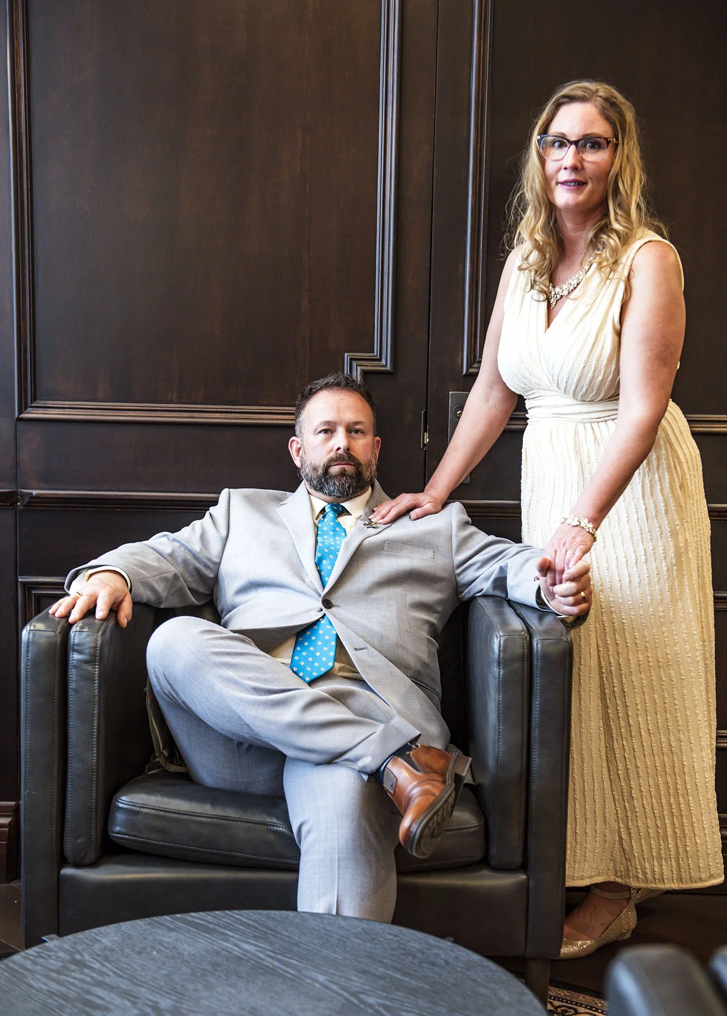 A man with a beard and a woman with glasses and curly blonde hair holding hands in an office setting. The man is seated in a black armchair, wearing a light gray suit, yellow shirt, and blue polka dot tie. The woman is standing beside him, wearing a 