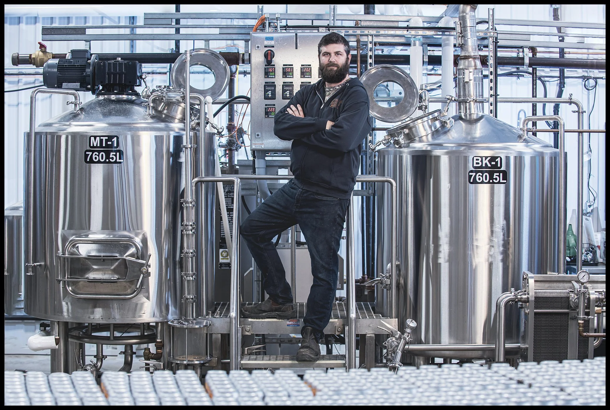 A man with a beard in a black hoodie and jeans is standing with crossed arms in front of large stainless steel industrial tanks with labels MT-1 and BK-1, in a bright, high-tech manufacturing facility.