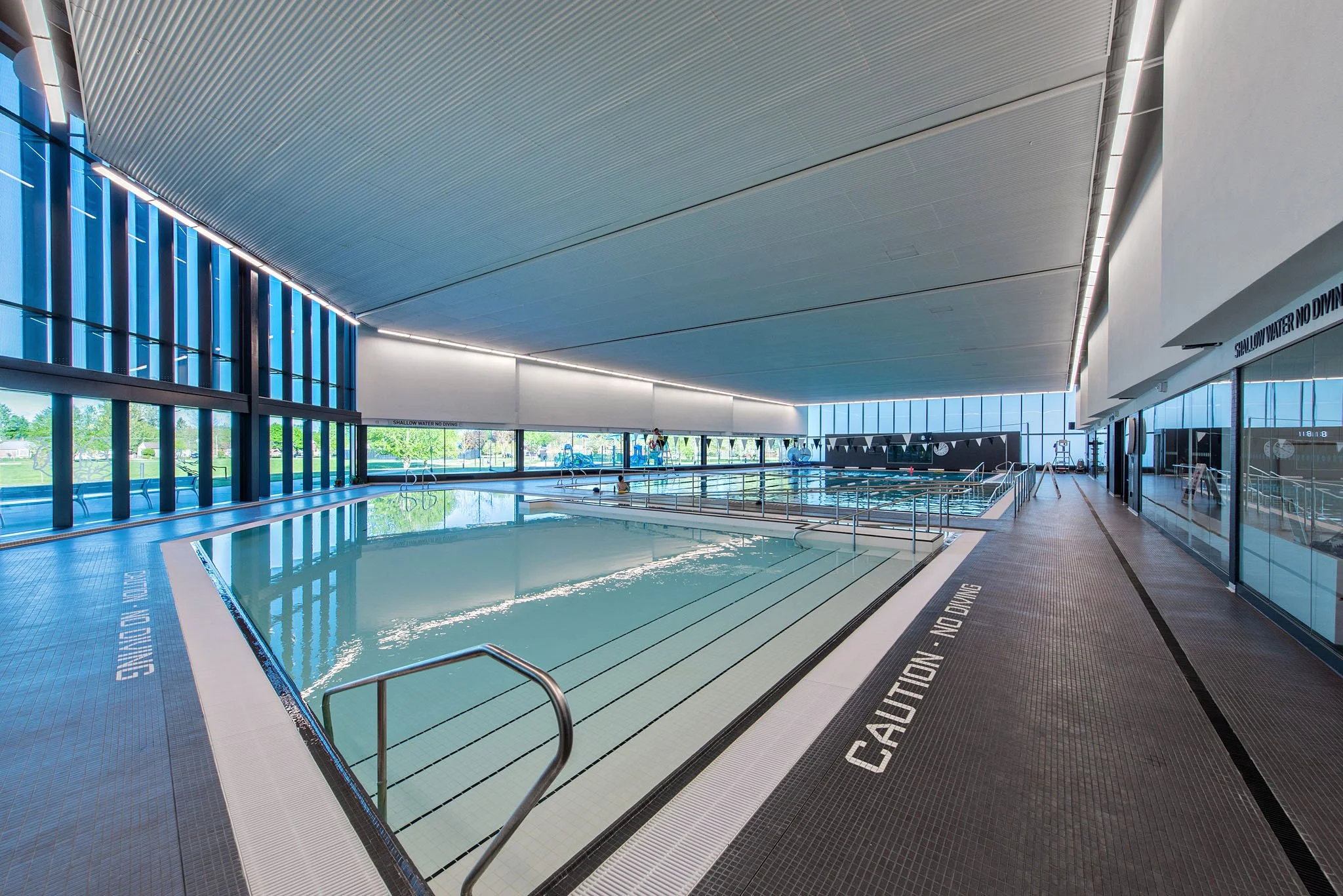 Indoor swimming pool with large windows, blue water, and modern architecture, with a sign on the floor indicating caution for shallow water.