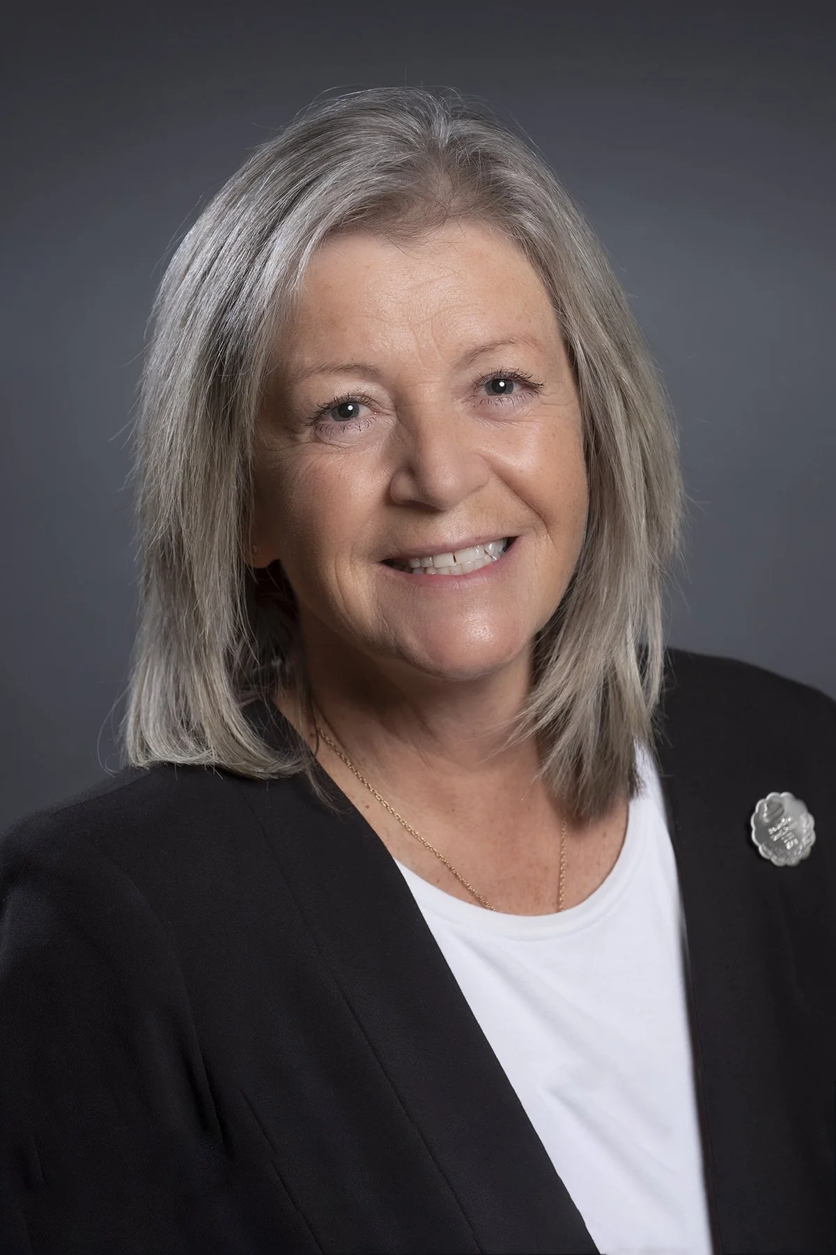 A middle-aged woman with shoulder-length gray hair, wearing a black blazer over a white shirt, smiling at the camera against a dark gray background.