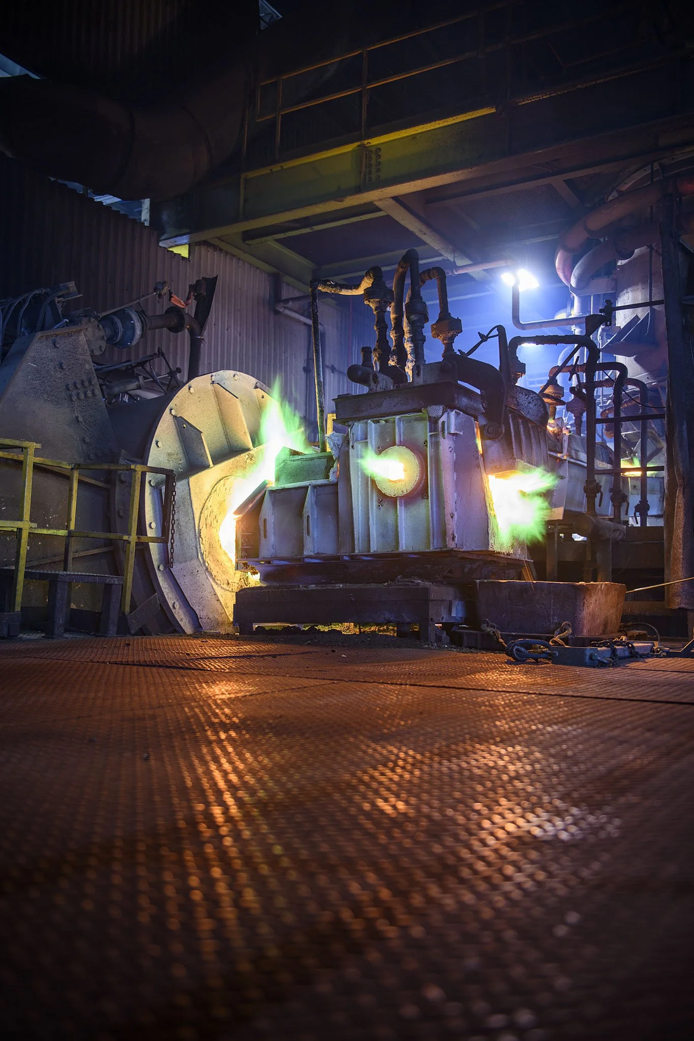 Industrial factory scene with molten metal pouring from a furnace, illuminated by bright sparks and green flames, surrounded by pipes and machinery.