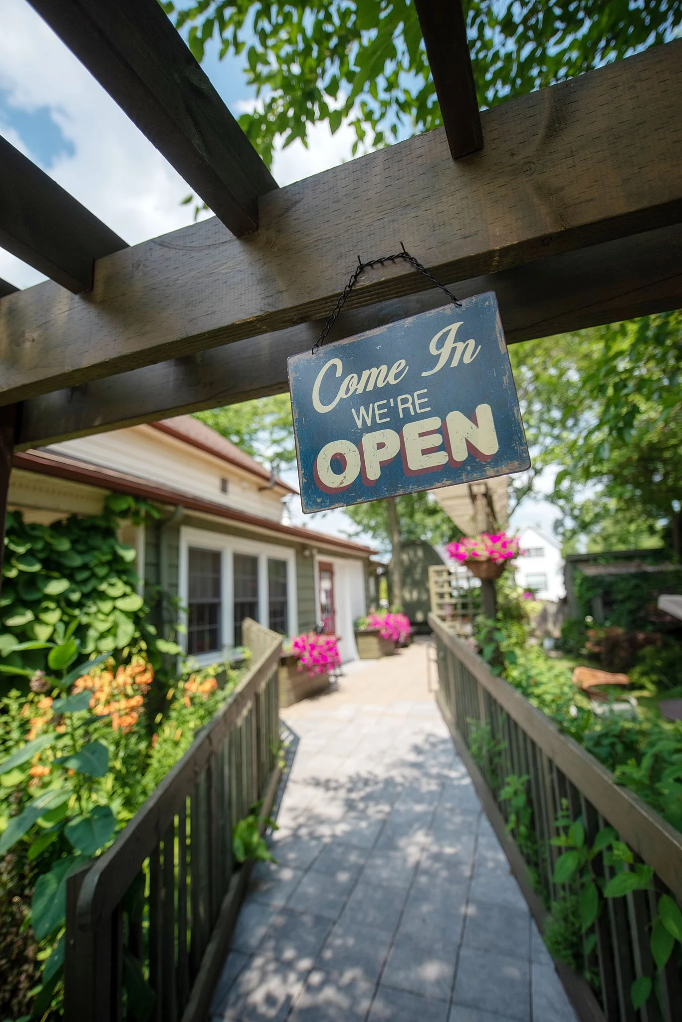 A pathway about to lead into a house with a sign hanging from the wooden beam above that says, 'Come In We're Open.' The pathway is flanked by garden beds filled with pink and orange flowers and lush green foliage. The house exterior features gray si