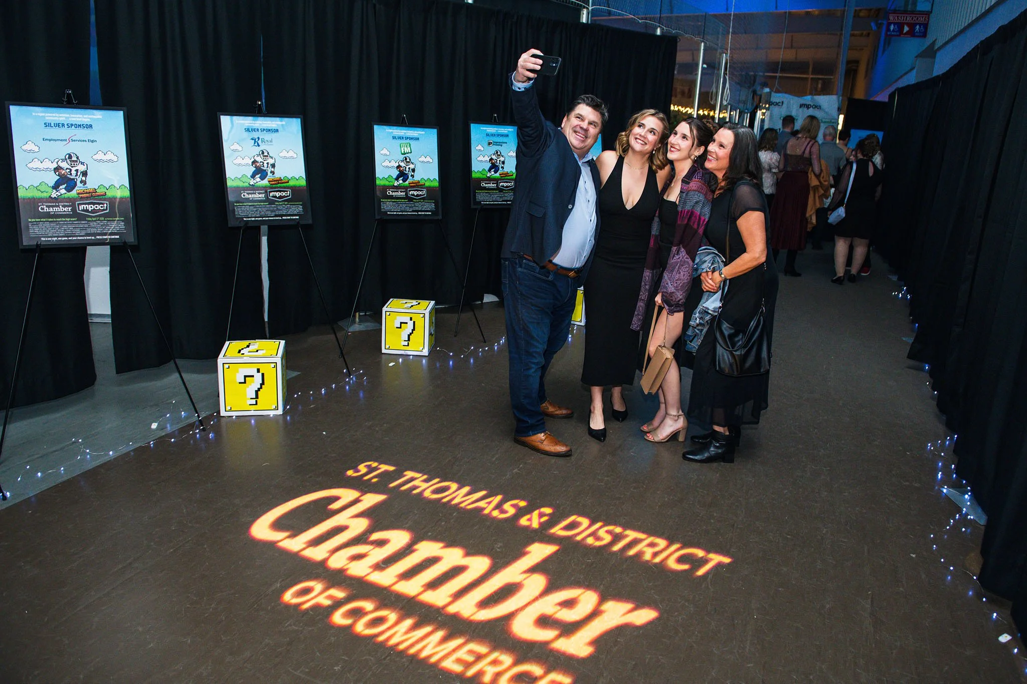 Four people taking a selfie together at an event with posters and decorations on black fabric backgrounds.