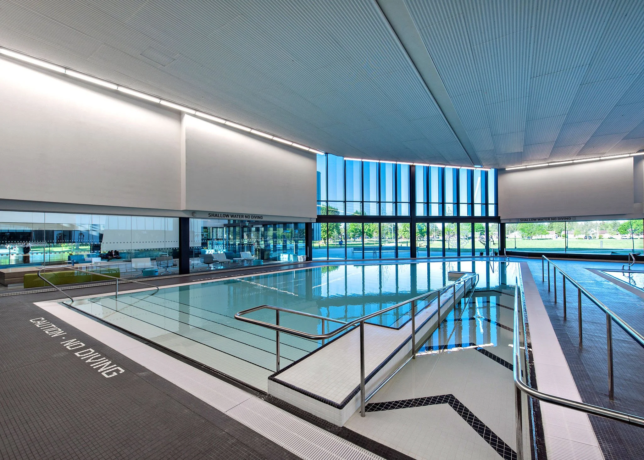 Indoor swimming pool area with large windows, pool, and seating area, with a sign that reads 'Shallow Water No Diving' on the poolside.