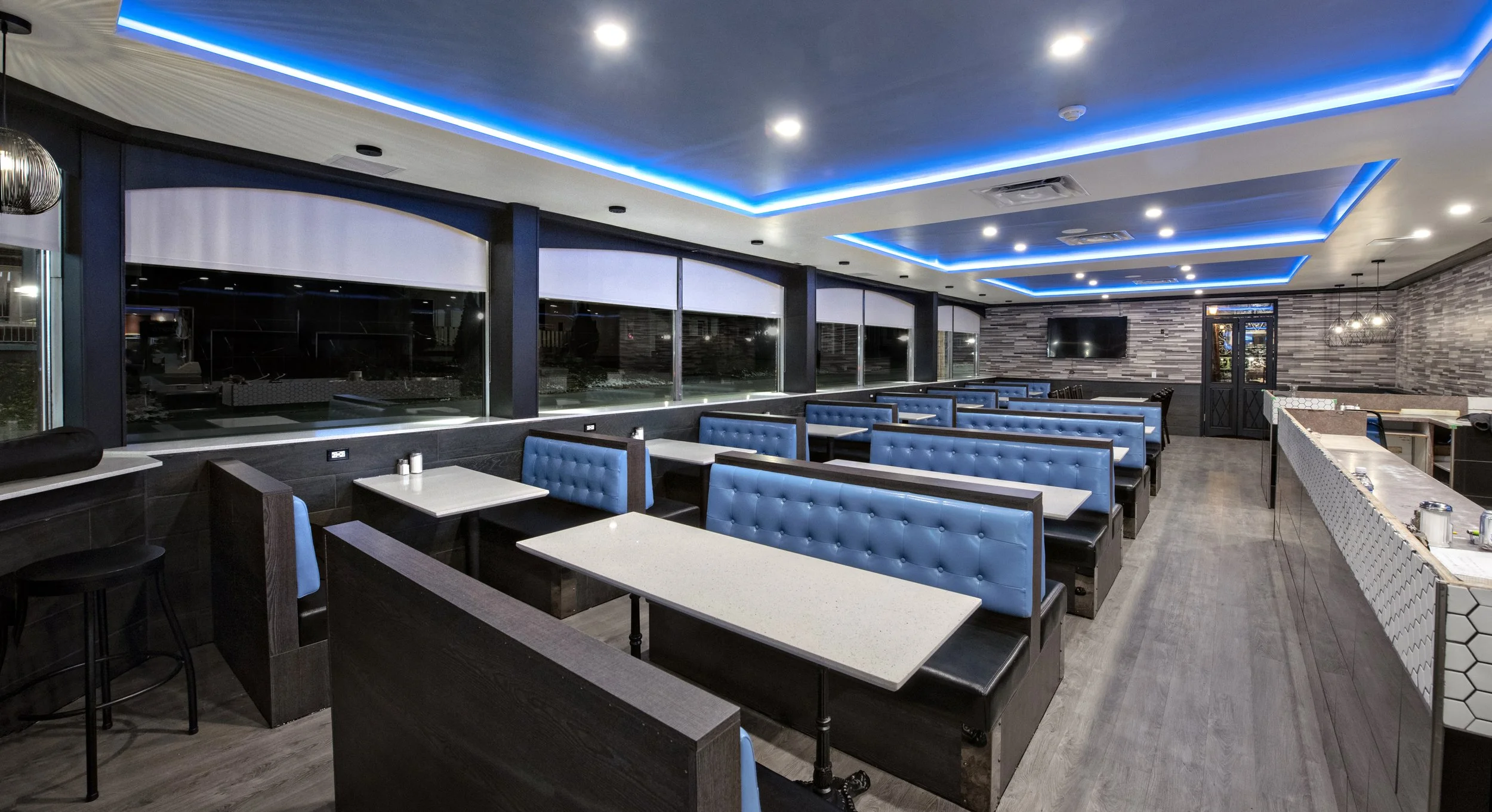 A modern restaurant interior with blue upholstered booths, white tables, large windows, and a gray tile accent wall, illuminated by ceiling lights and blue LED strip lighting.
