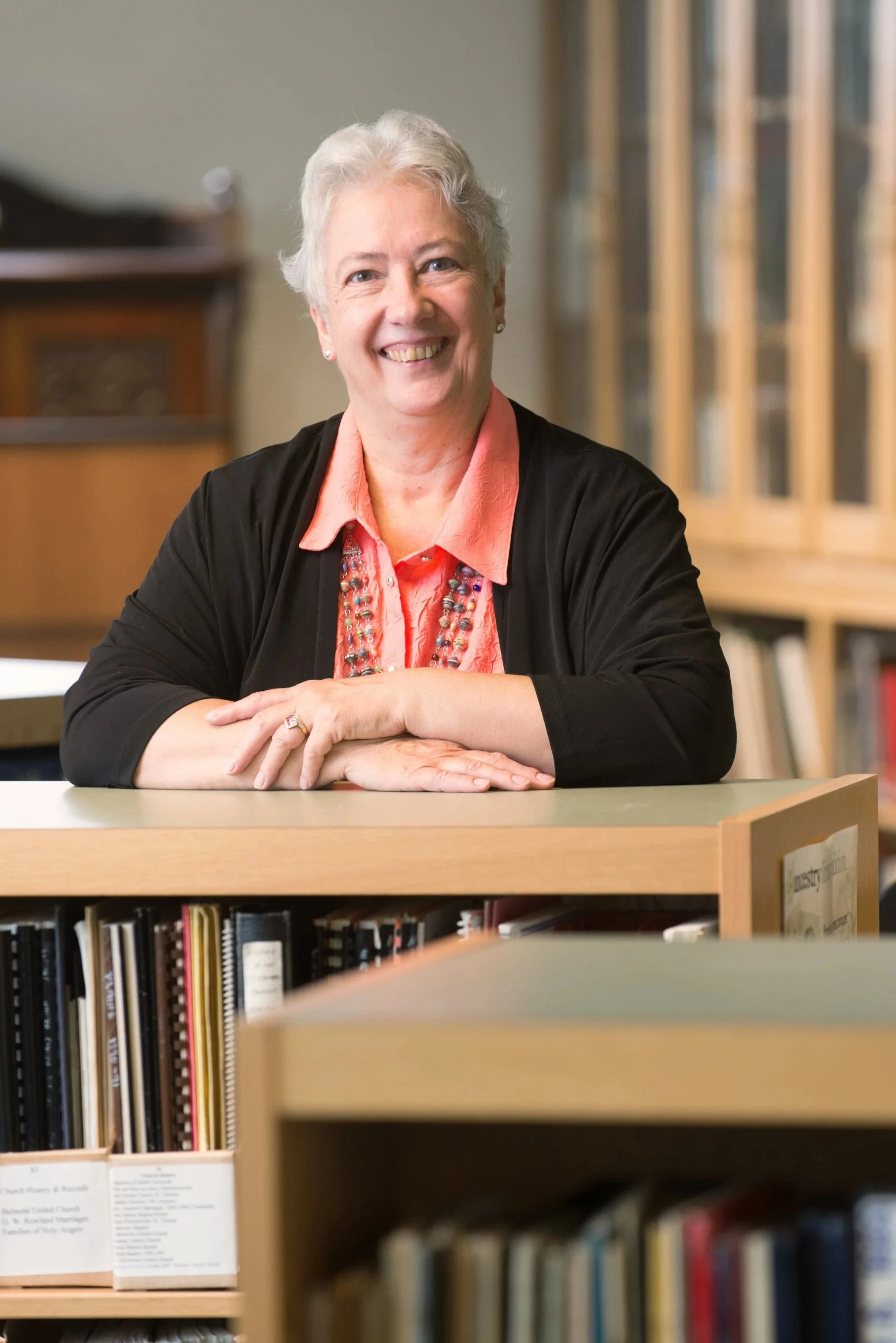 A smiling elderly woman with short gray hair sitting at a desk in a library, with bookshelves in the background.