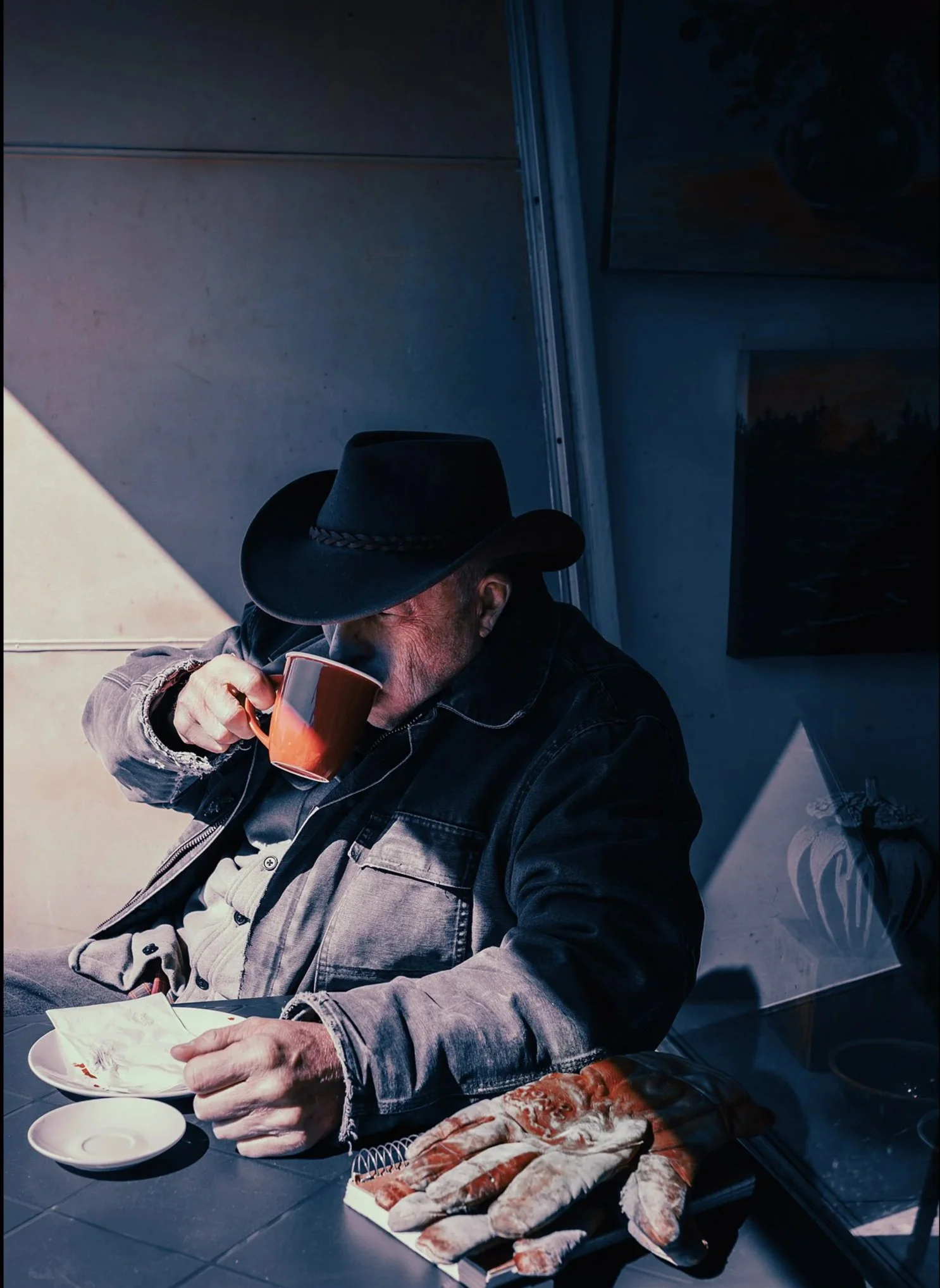 A man with a fedora hat and plaid jacket sitting at a table, drinking from a mug, with fish on a tray in front of him.