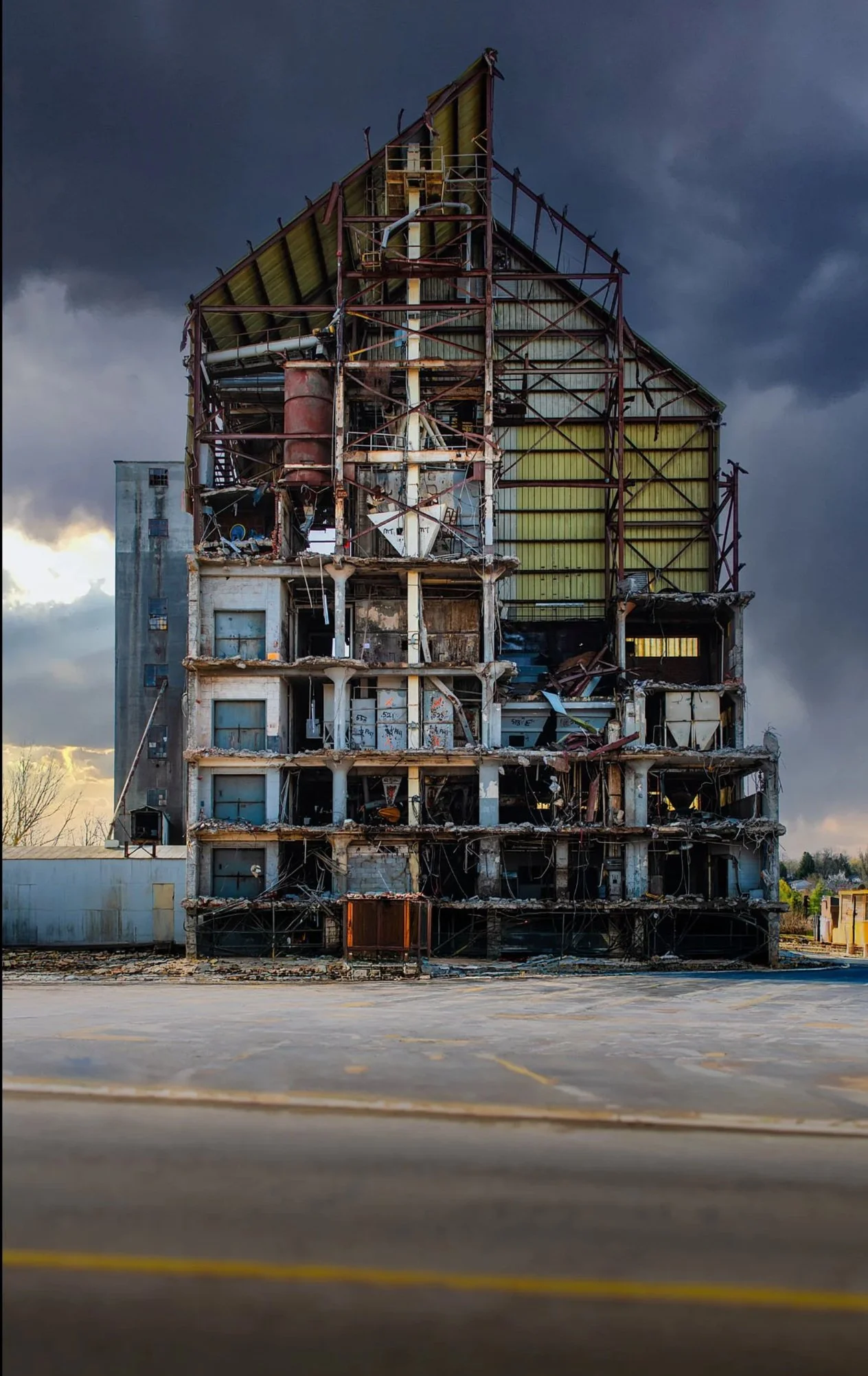 Dilapidated multi-story building with exposed structural elements, surrounded by an empty parking lot under a dark stormy sky.
