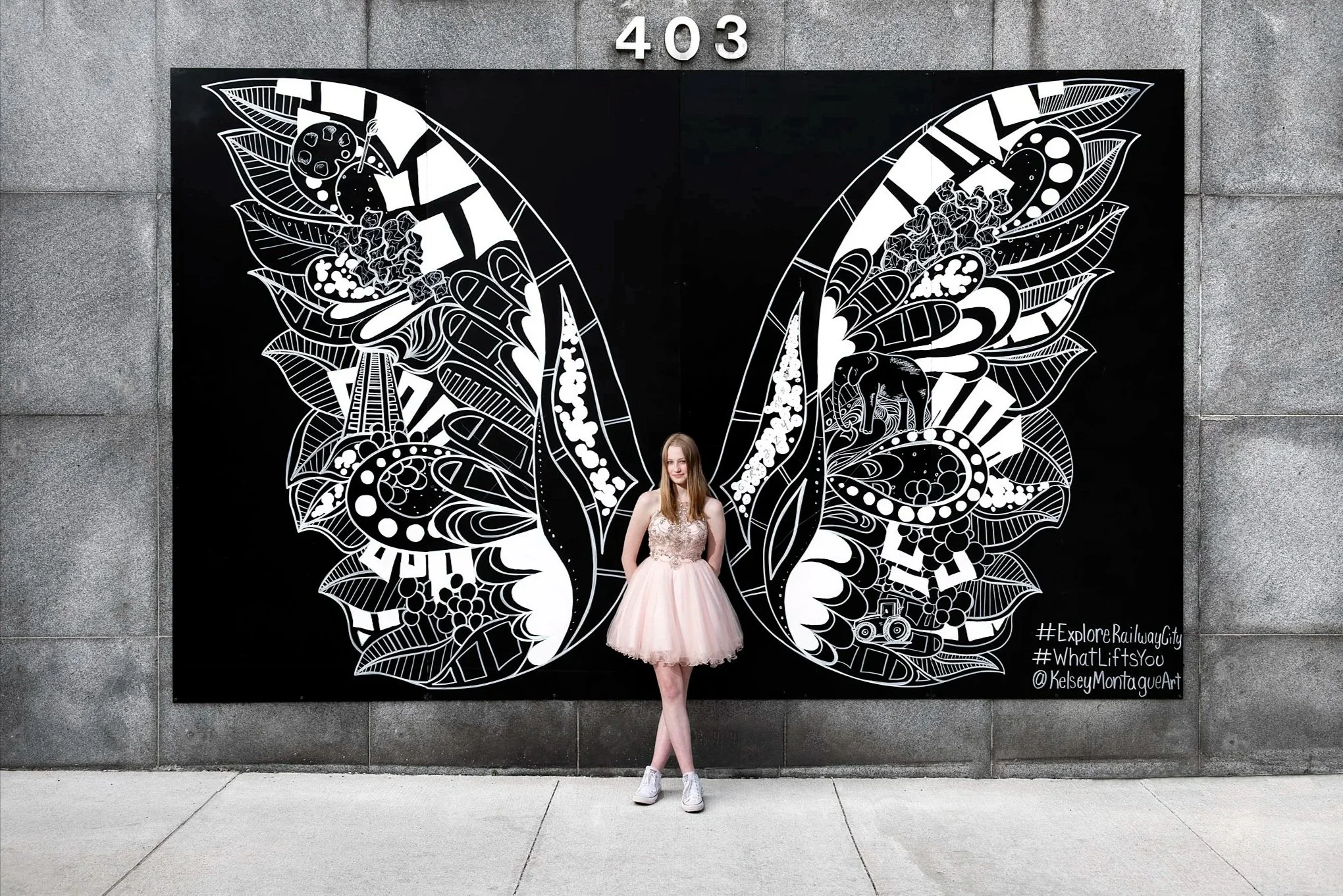 A woman in a light pink dress and sneakers standing in front of a large black and white butterfly mural on a building wall, with the numbers 403 above.
