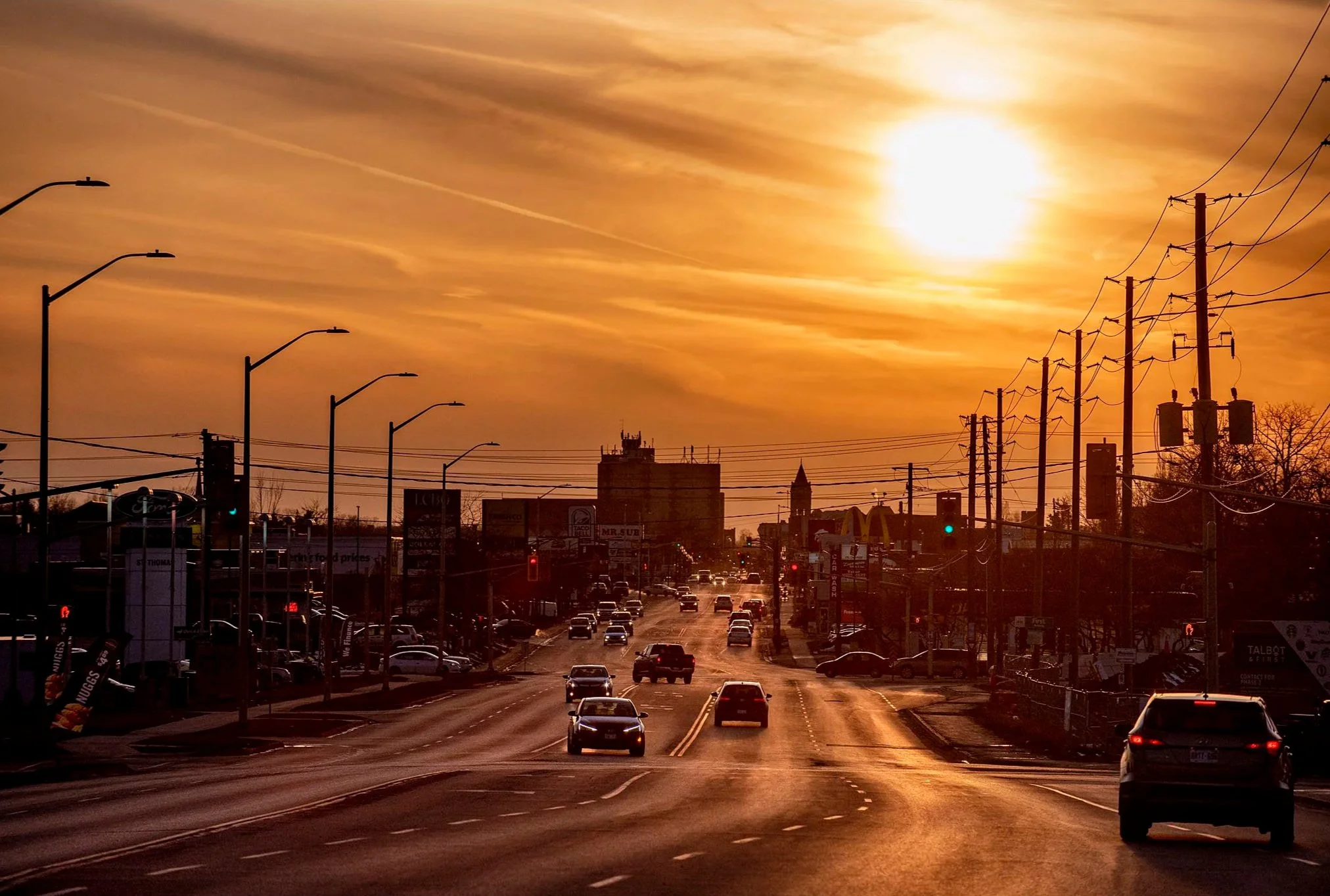 A busy city street during sunset with cars driving on the road, power lines and streetlights along the sides, and buildings in the background. The sky is orange with the sun visible, creating a warm glow.