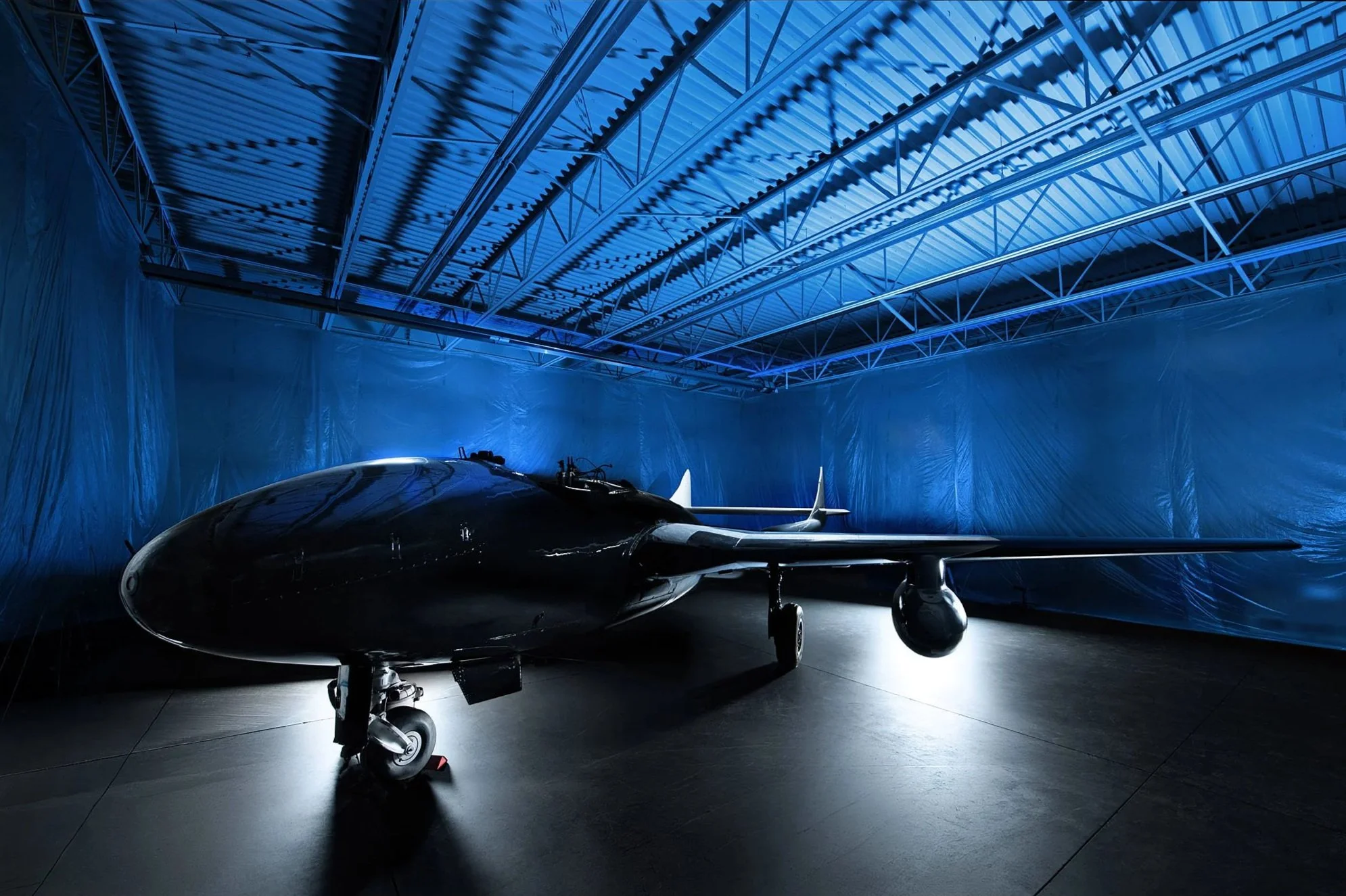 A sleek black fighter jet aircraft inside a blue-lit hangar with a metallic ceiling structure.