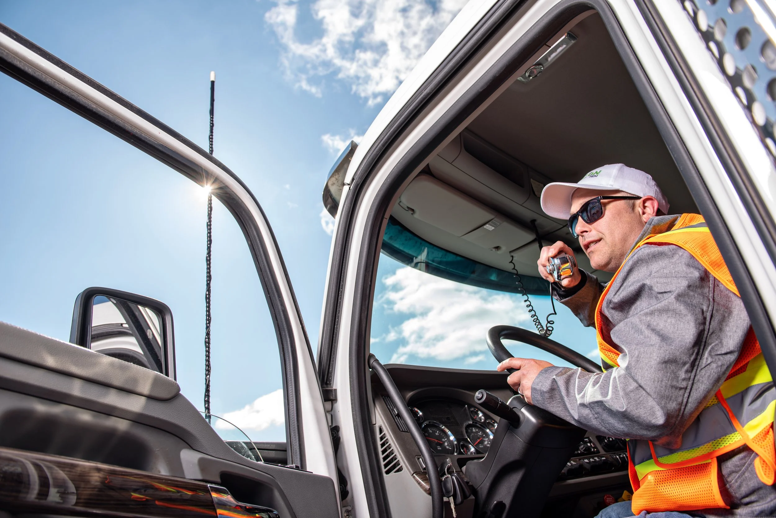 A man wearing sunglasses, a white cap, and a reflective safety vest sitting in the driver's seat of a large truck, holding a radio microphone, with the door open, under a blue sky with some clouds.