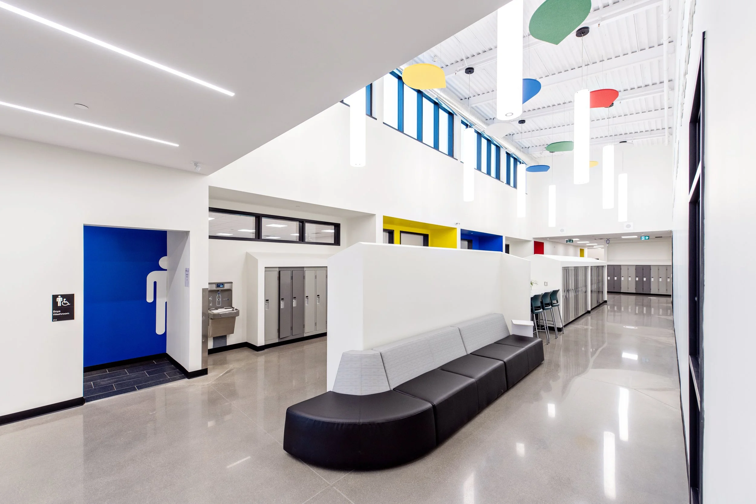 Modern school or public building interior with white walls, gray flooring, black benches and colorful wall features, including yellow, red, and blue accents, fluorescent ceiling lights, and a high ceiling with skylights.