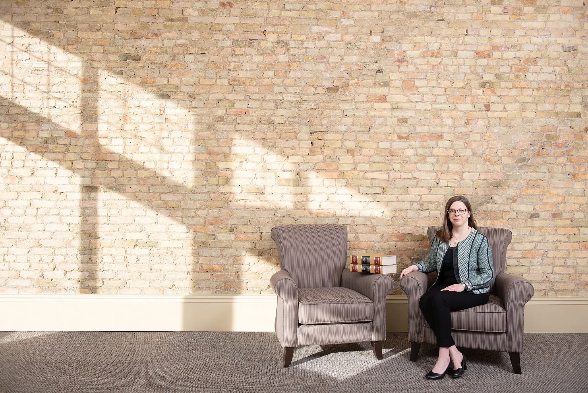 A woman sitting on a purple armchair with a stack of books on the armrest, in front of a brick wall with sunlight and shadow patterns. She is wearing glasses, a patterned blazer, and black pants.