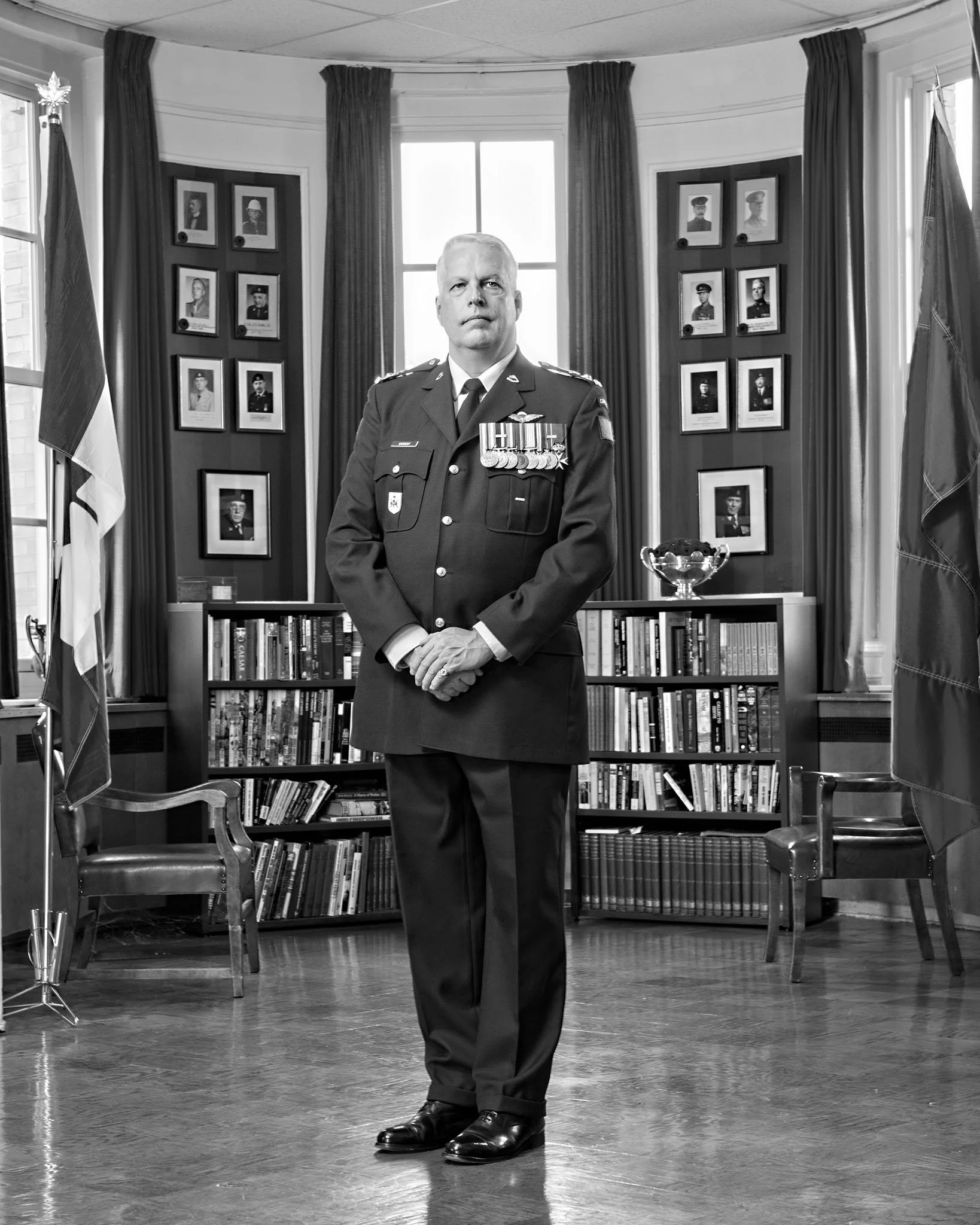 A man in a military uniform with medals and ribbons standing in a room with framed photographs on the wall behind him, with flags on either side of the photo. The room has large windows with curtains and a bookshelf.
