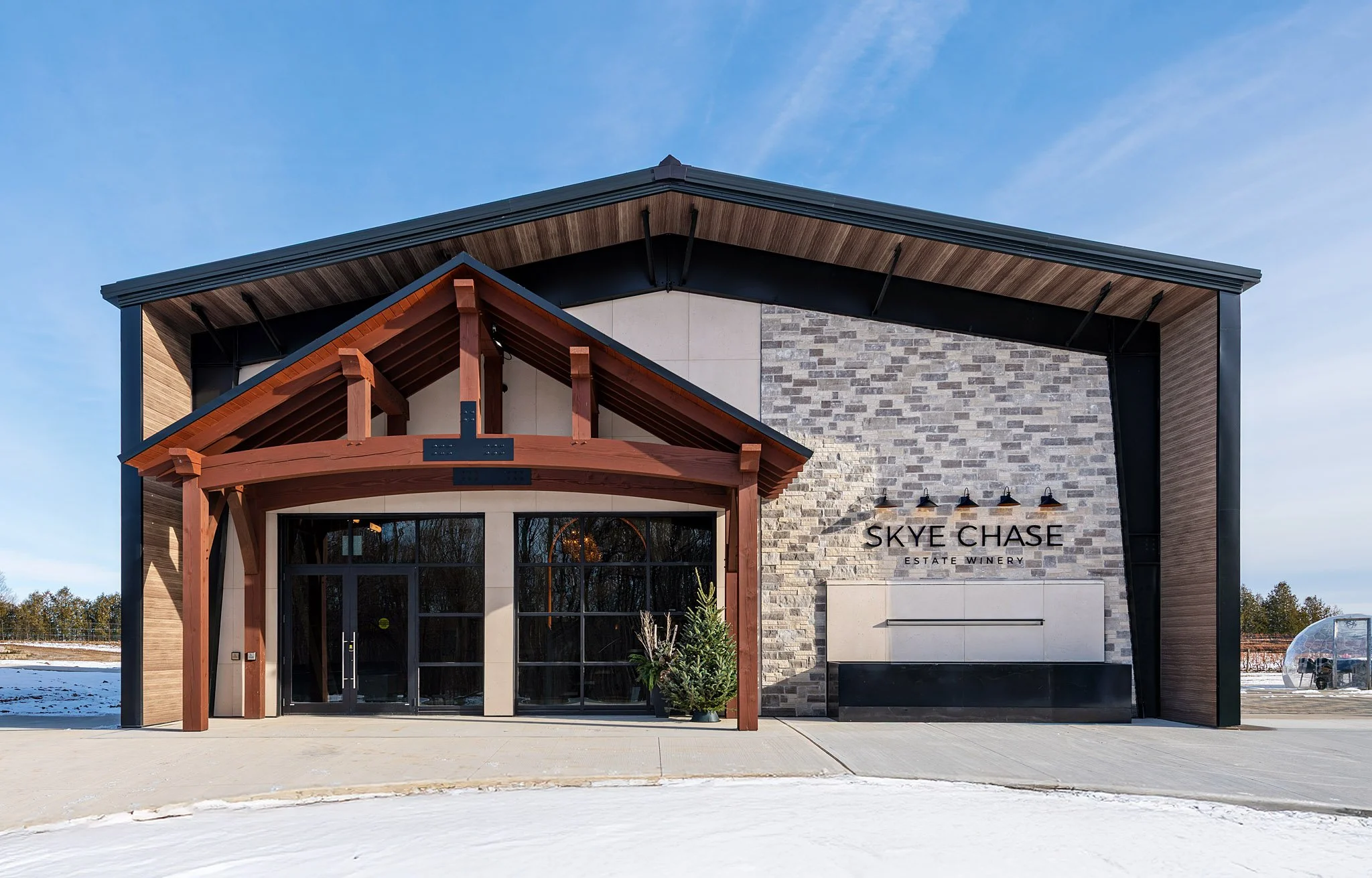 Modern building with a sign that reads 'Skye Chase Estate Winery,' featuring a stone facade, wooden accents, and large glass doors, under a clear blue sky.