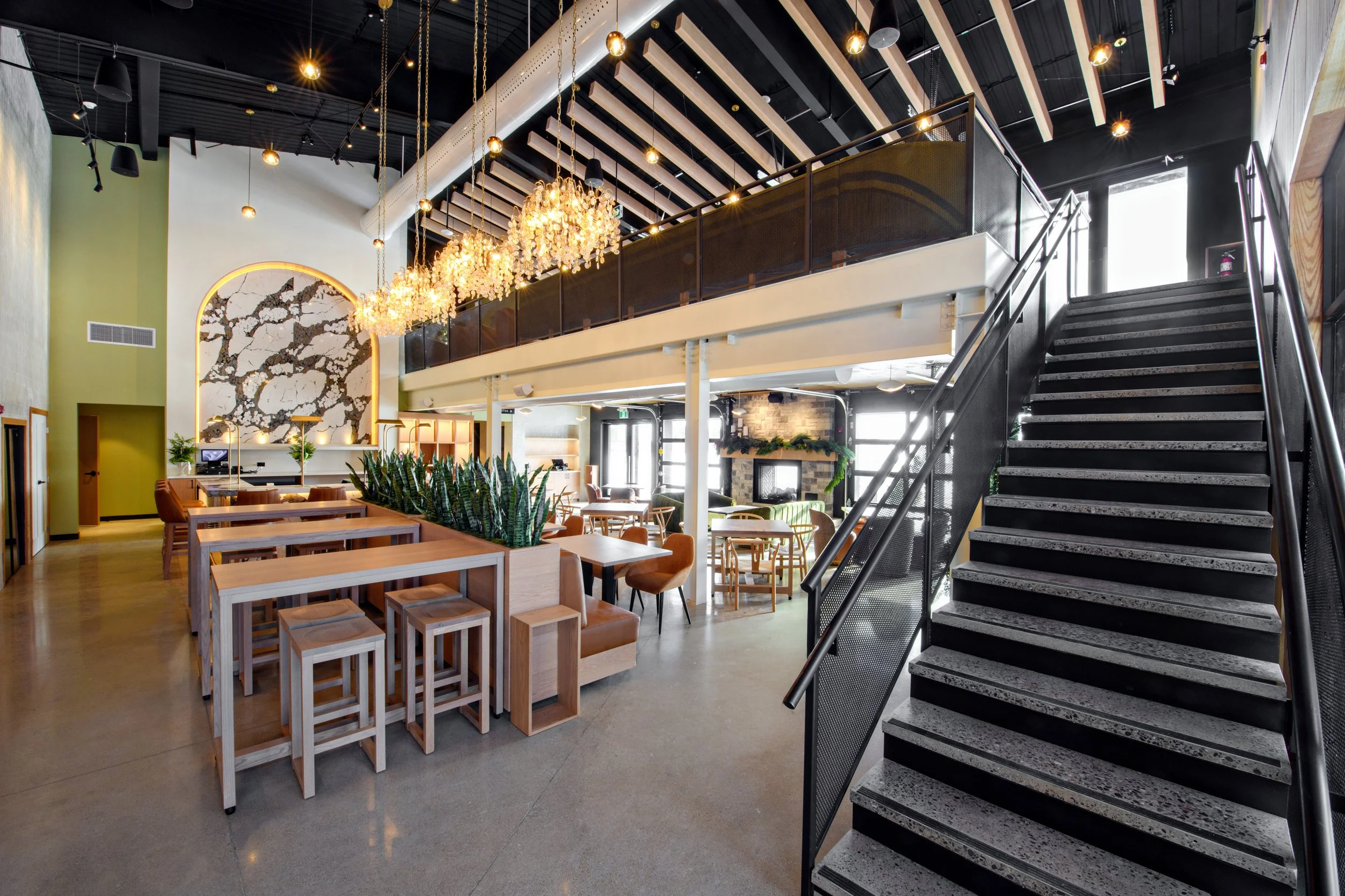 Interior of a modern restaurant or cafe with high ceilings, hanging light fixtures, and a staircase. Wooden furniture and a decorative wall art piece are visible.