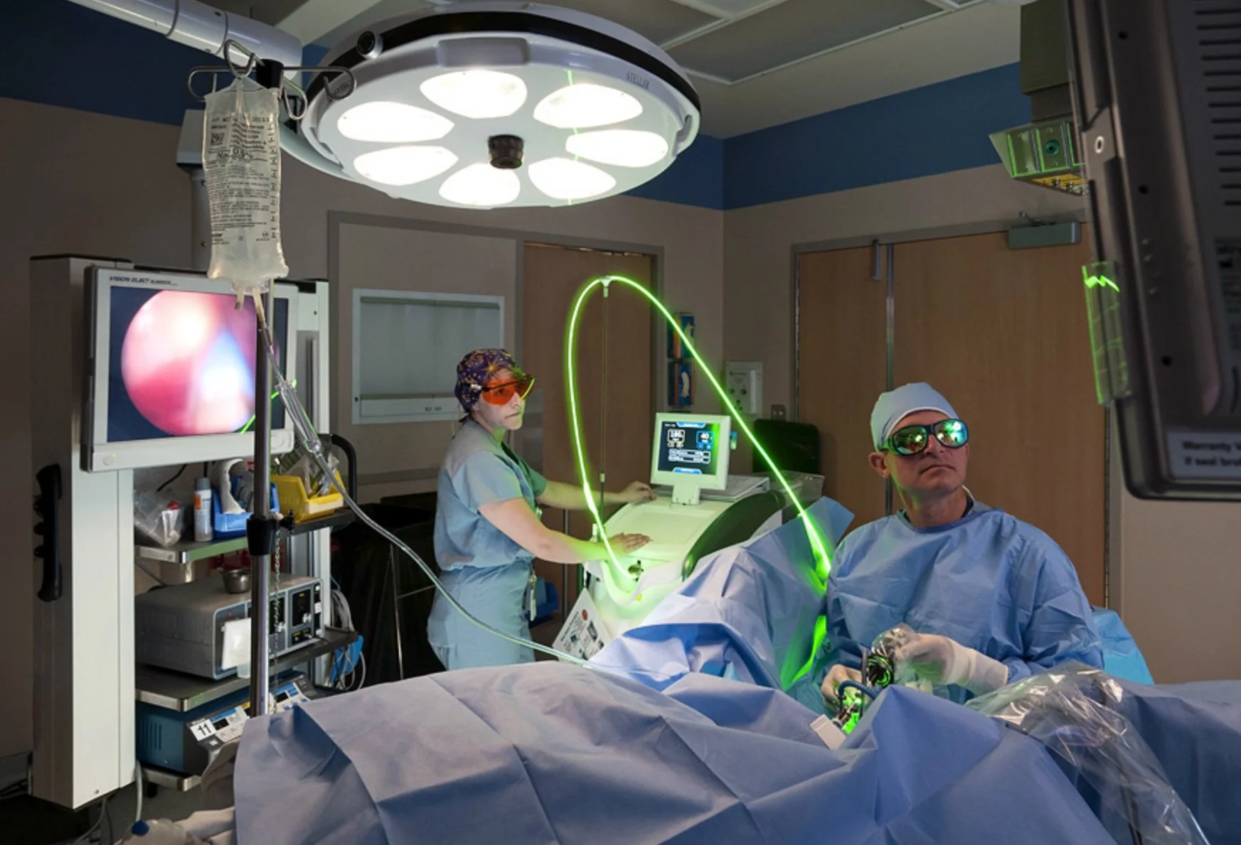 Medical professionals in an operating room performing surgery with advanced technology, including a monitor showing an endoscopy image and green lasers.