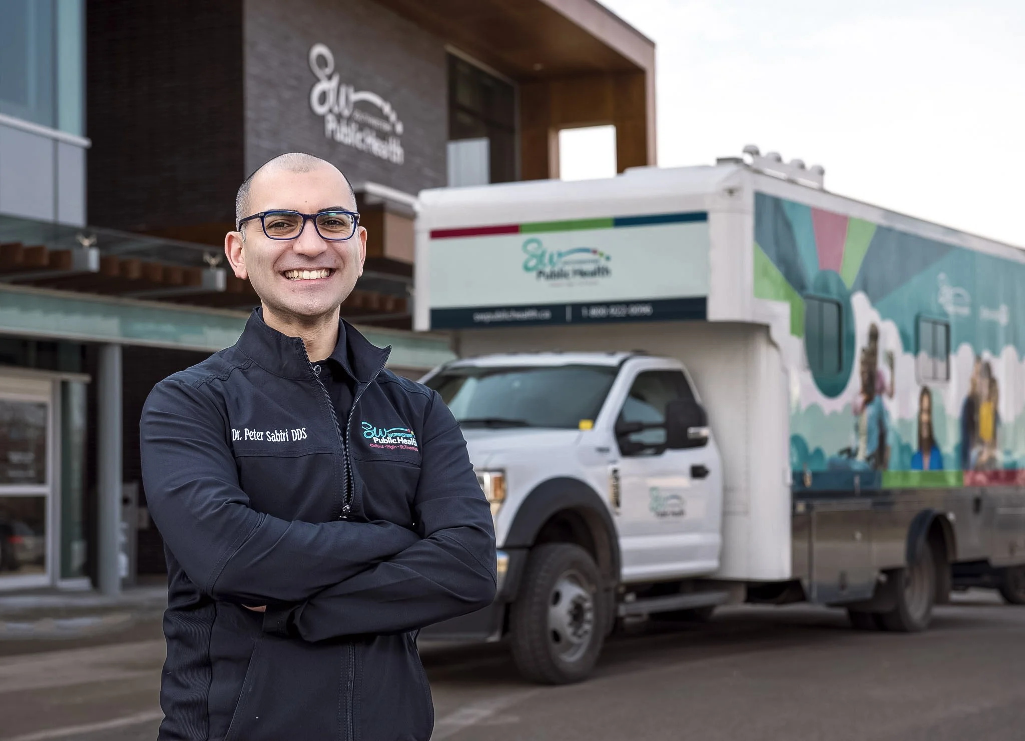 A smiling man with glasses and a black jacket standing outdoors in front of a mobile health clinic truck, with a building behind him that has a sign reading "Public Health". Executive portrait photography. 