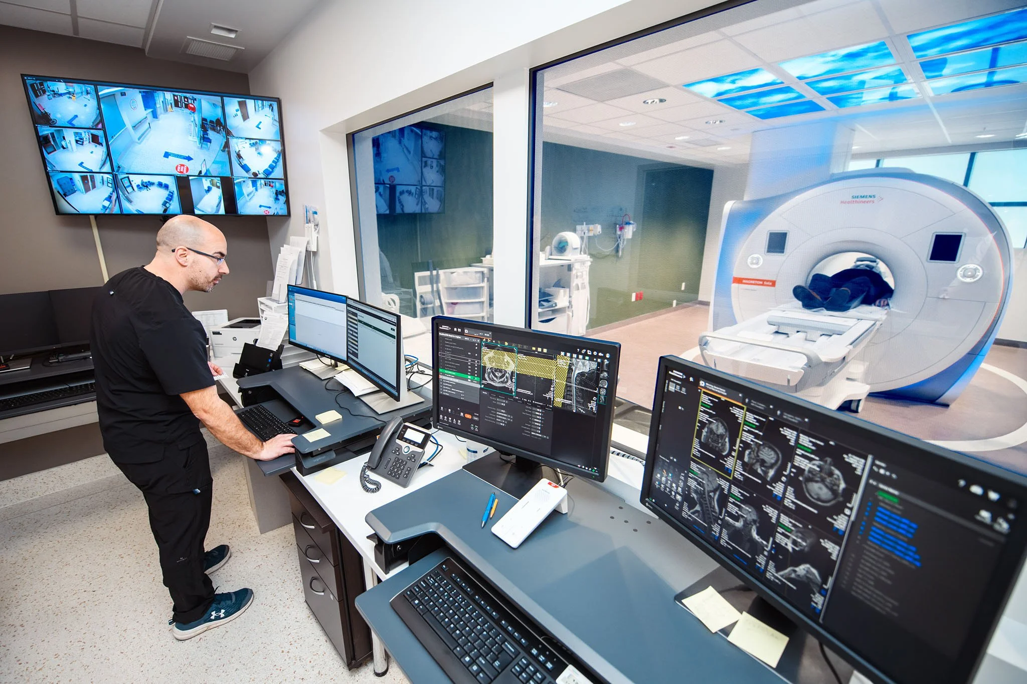 A man working at a control desk with multiple computer monitors and a large MRI machine in a hospital imaging room, with additional monitors on the wall showing various surveillance camera feeds of the hallway.