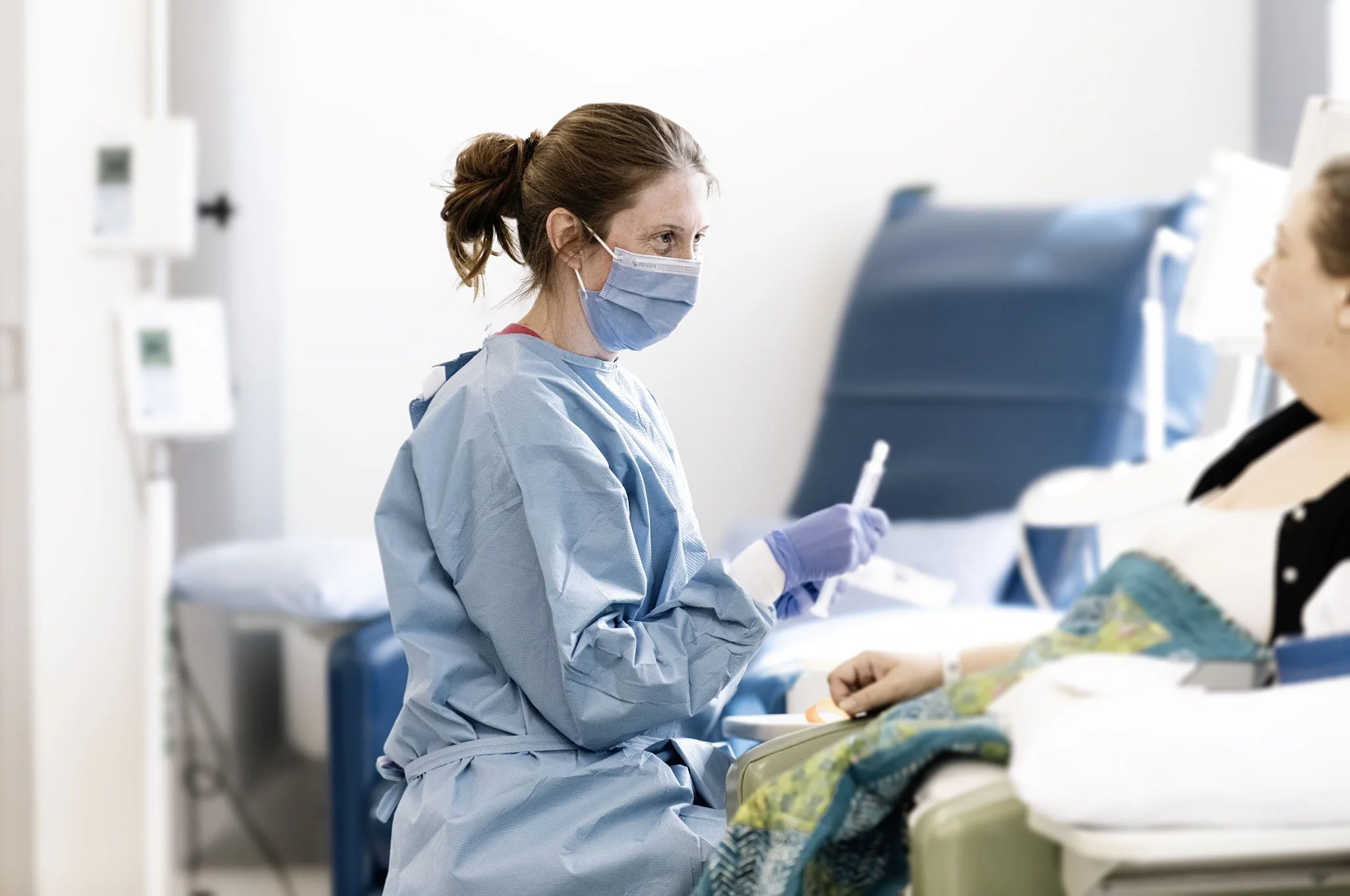 A healthcare professional wearing a blue surgical mask, gloves, and scrubs, interacting with a patient in a hospital or clinic setting.