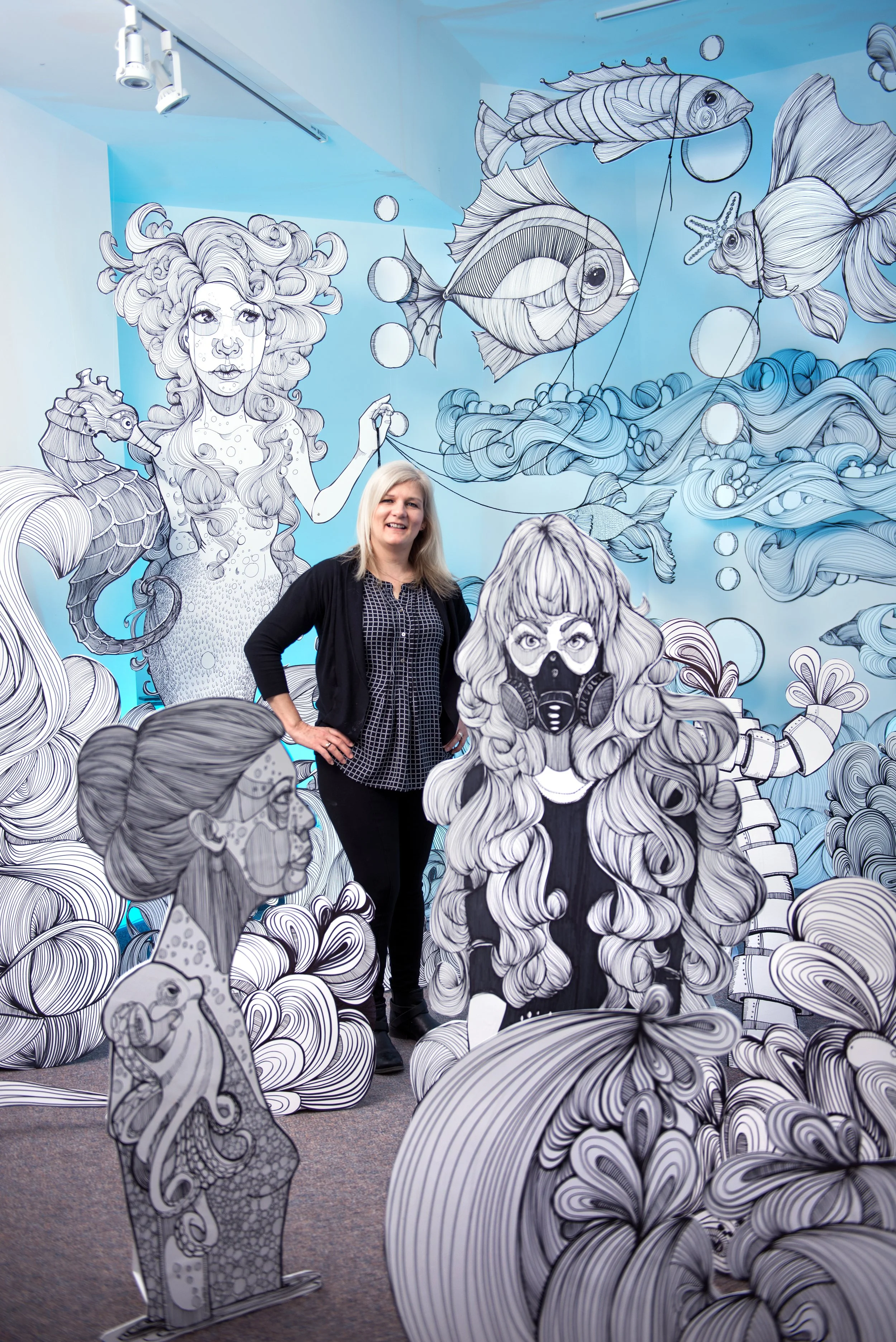 A woman with blonde hair smiling inside an art installation with black and white drawings of mermaids, fish, and sea creatures on the walls and life-sized cutouts of mermaids and sea animals. Executive portrait photography. 
