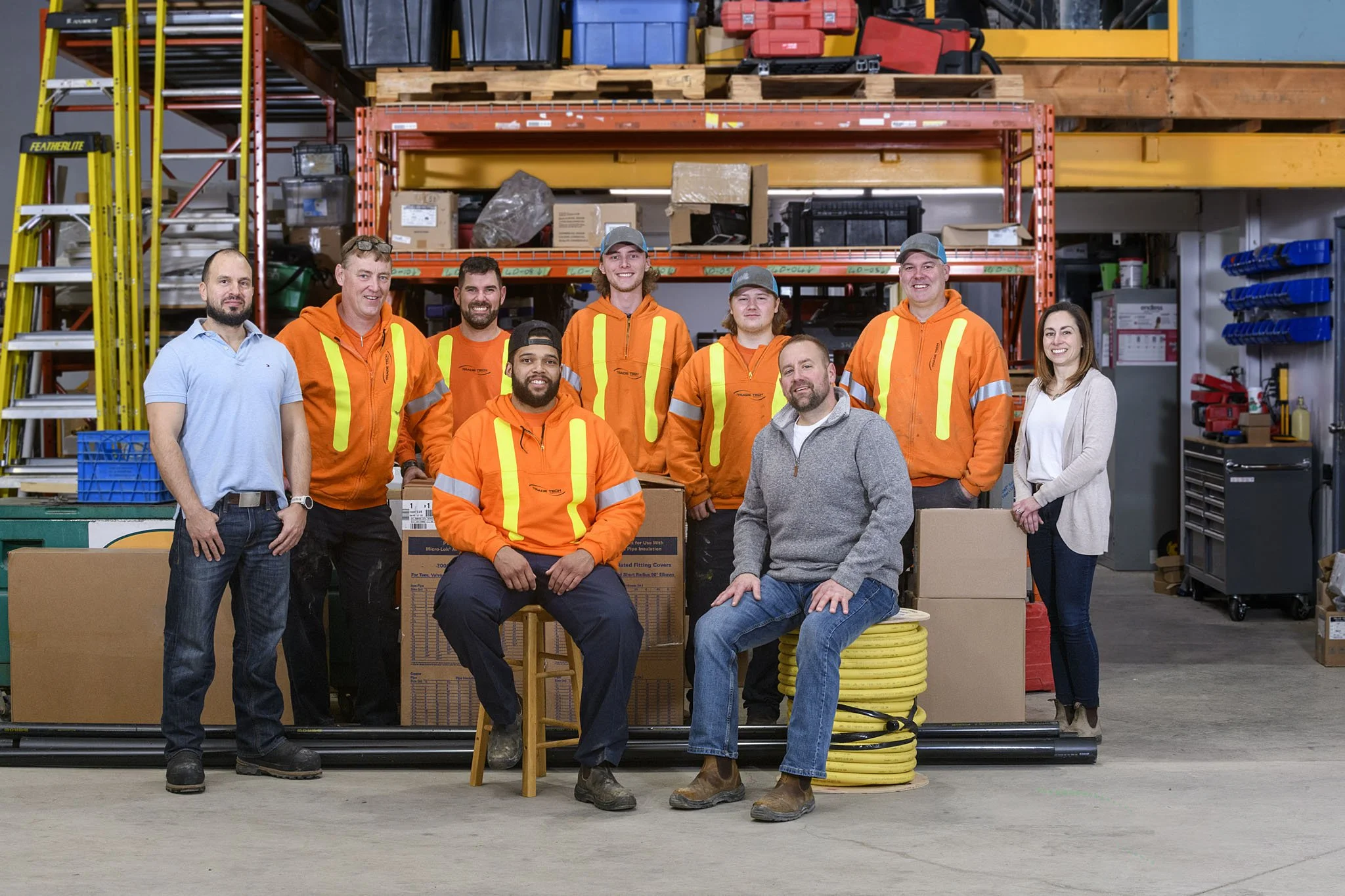 A group of nine people, including workers in orange safety jackets and casual attire, standing and sitting in a warehouse with shelves filled with boxes, tools, and industrial equipment.