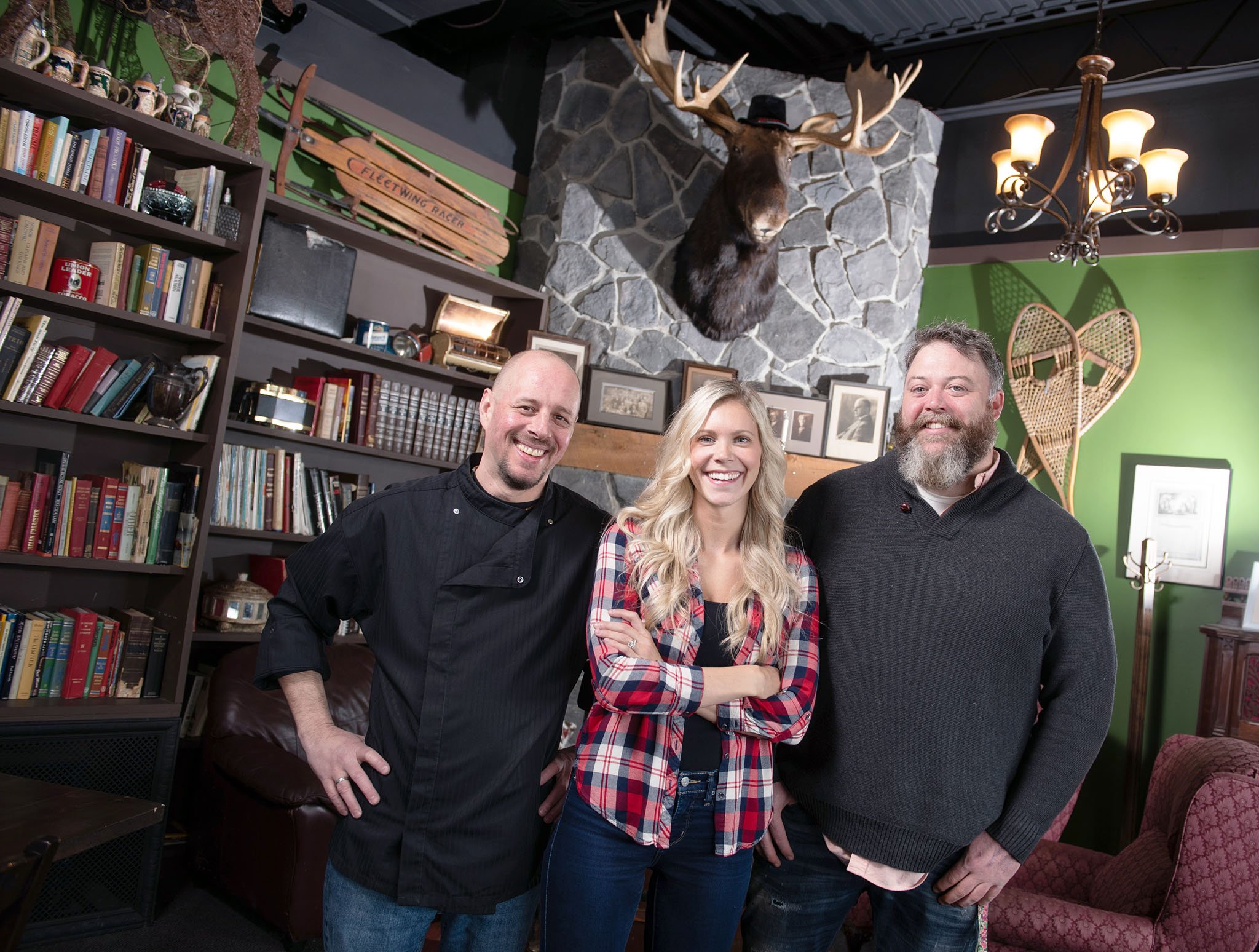 A group of three smiling people standing together in a cozy room with bookshelves, framed photos, and vintage decor, including a mounted elk head on a stone fireplace. They are casually dressed, and the room has warm lighting and a rustic atmosphere.