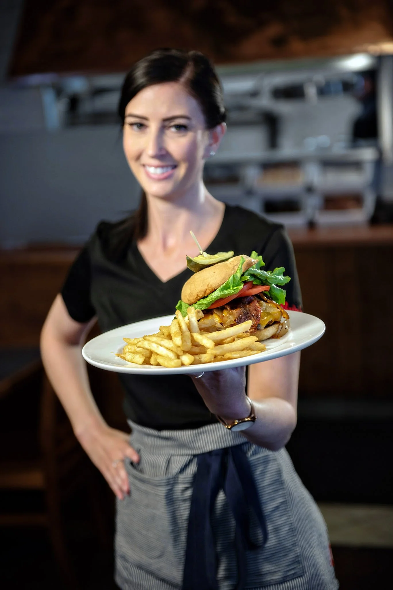 A woman with dark hair, wearing a black top and striped gray apron, presents a plate of French fries and a burger with lettuce, tomato, pickles, and bacon, in a restaurant setting. Commercial photography 
