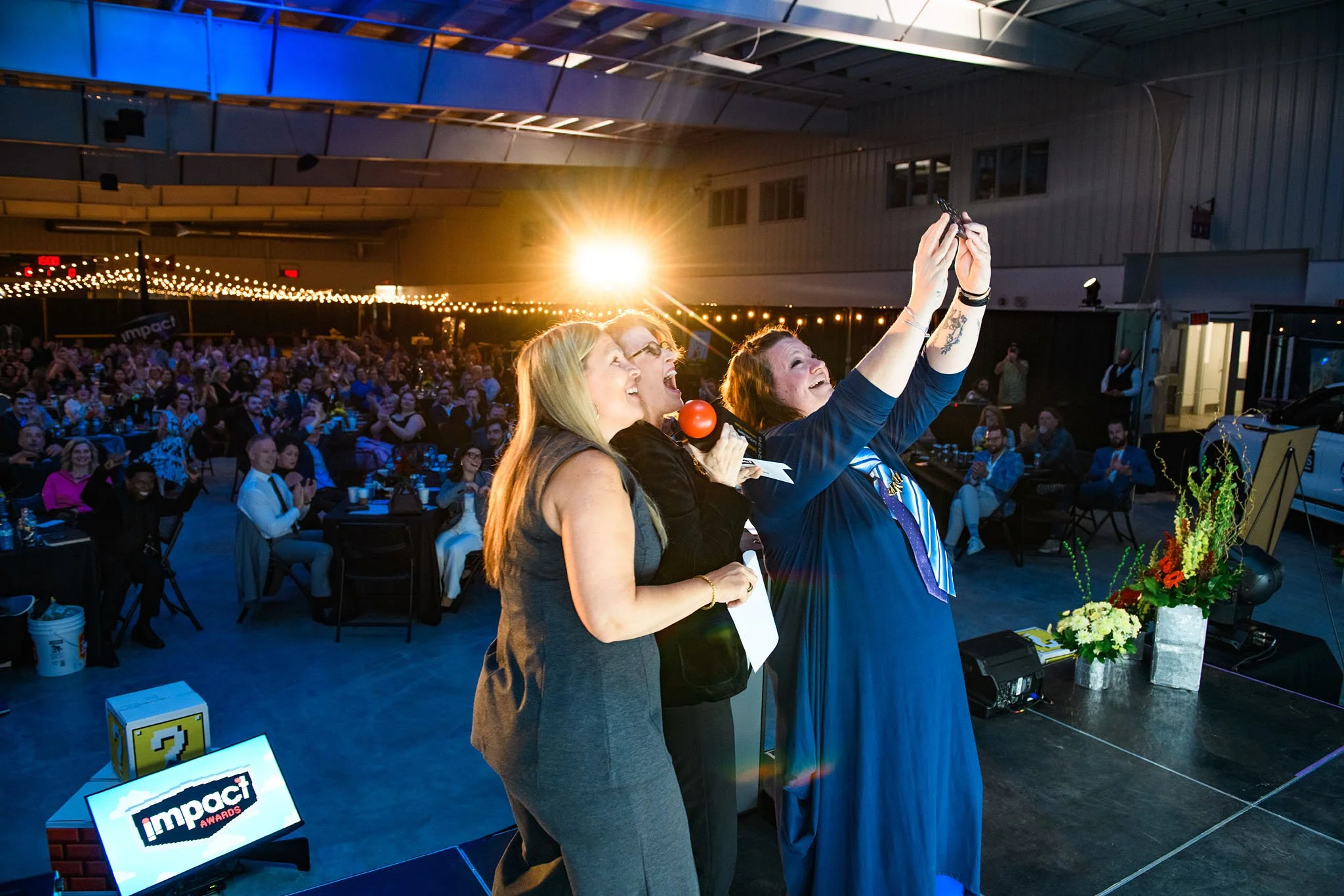 Three women taking a selfie at an award event, with a large audience in the background and decorated inside a large venue.