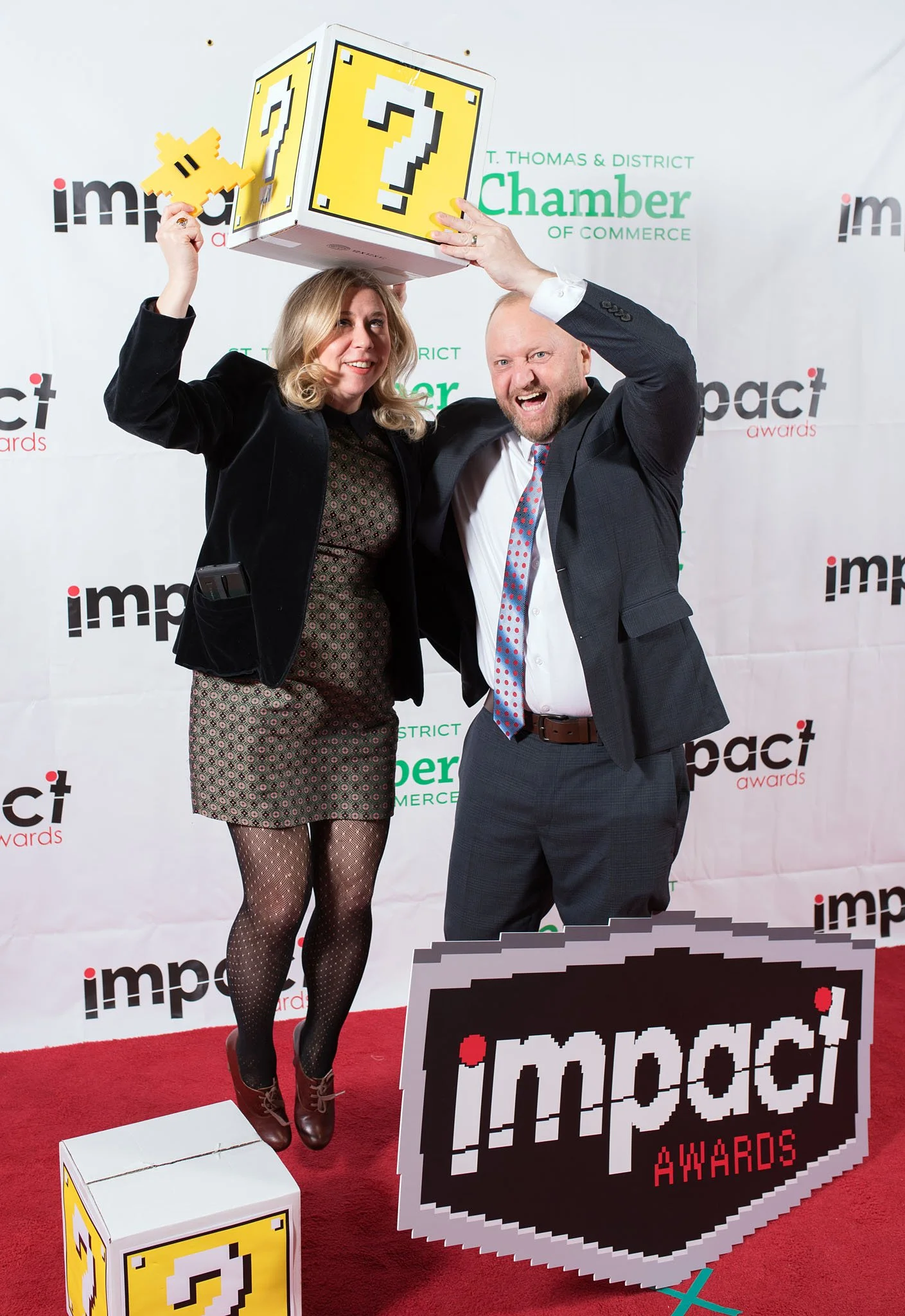 Two people, a woman in a black blazer and patterned dress, and a man in a dark suit with a polka dot tie, are celebrating at the Impact Awards. The woman is holding a pixelated yellow question mark sign, and the man is holding a pixelated yellow ques