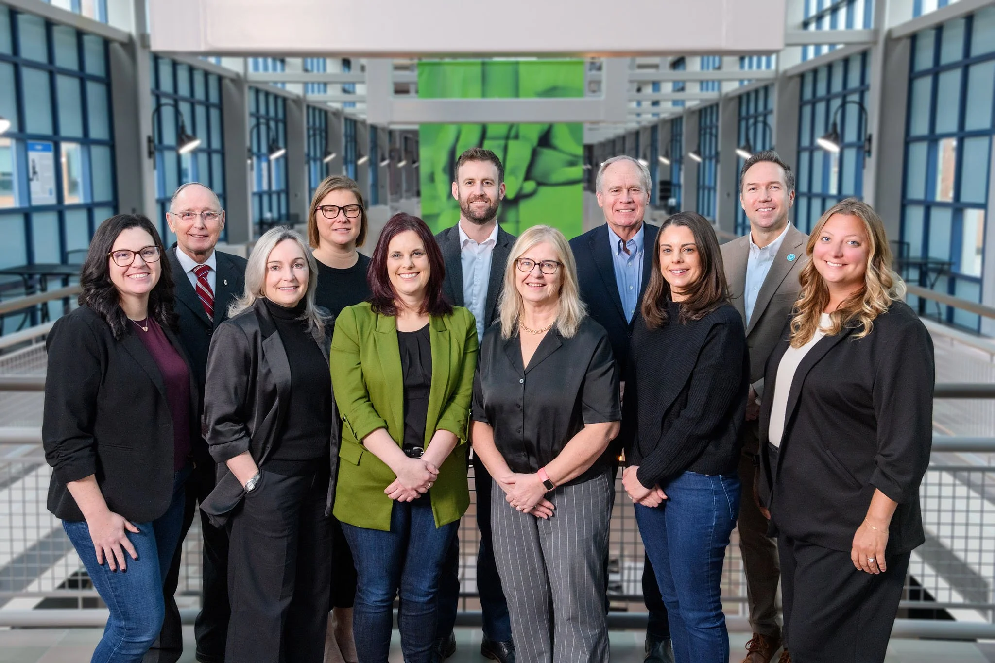 A diverse group of twelve professionals standing together indoors in a modern building with glass walls and metal railings, smiling at the camera.