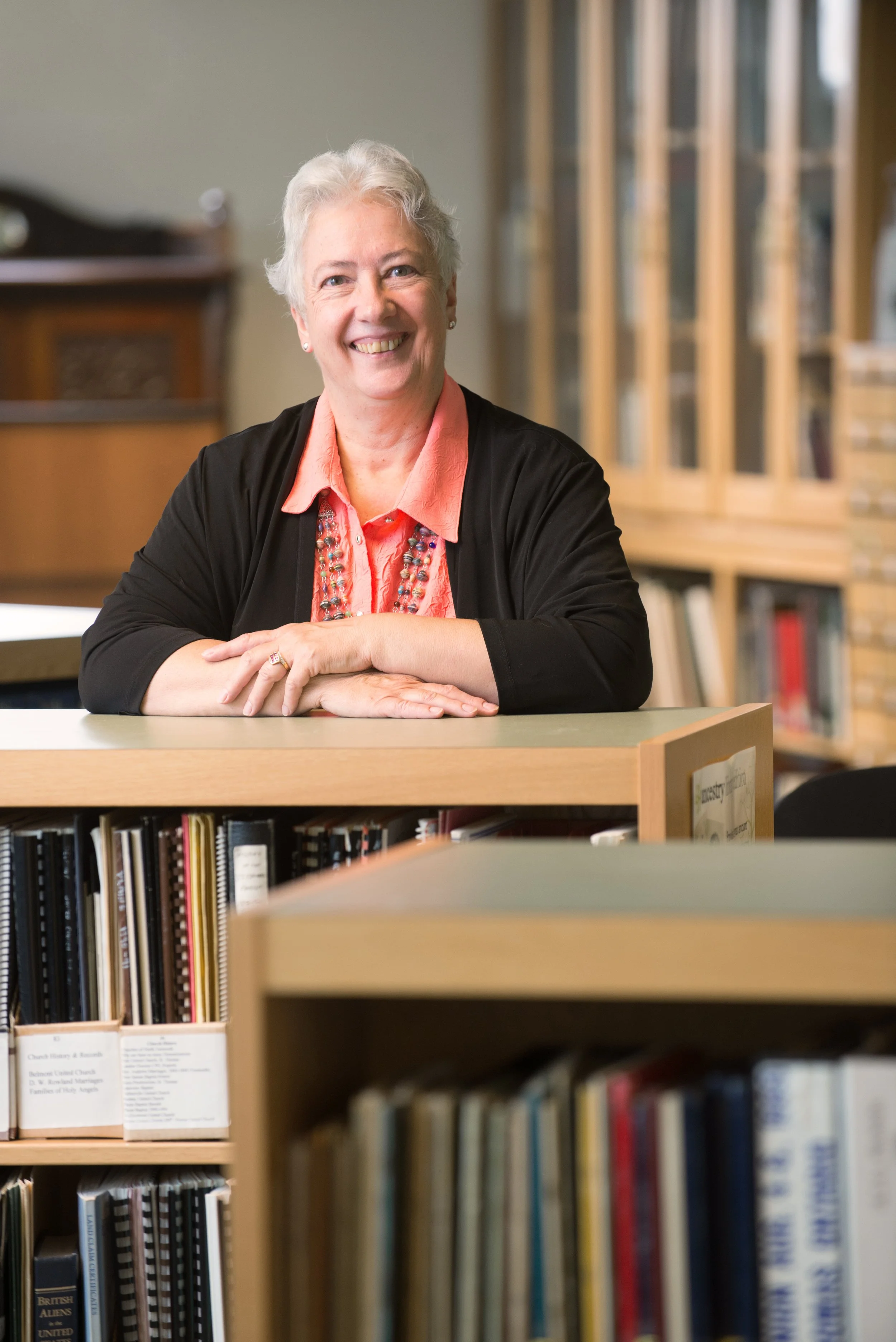 A smiling elderly woman with short gray hair sitting at a desk in a library, with bookshelves filled with books in the background.