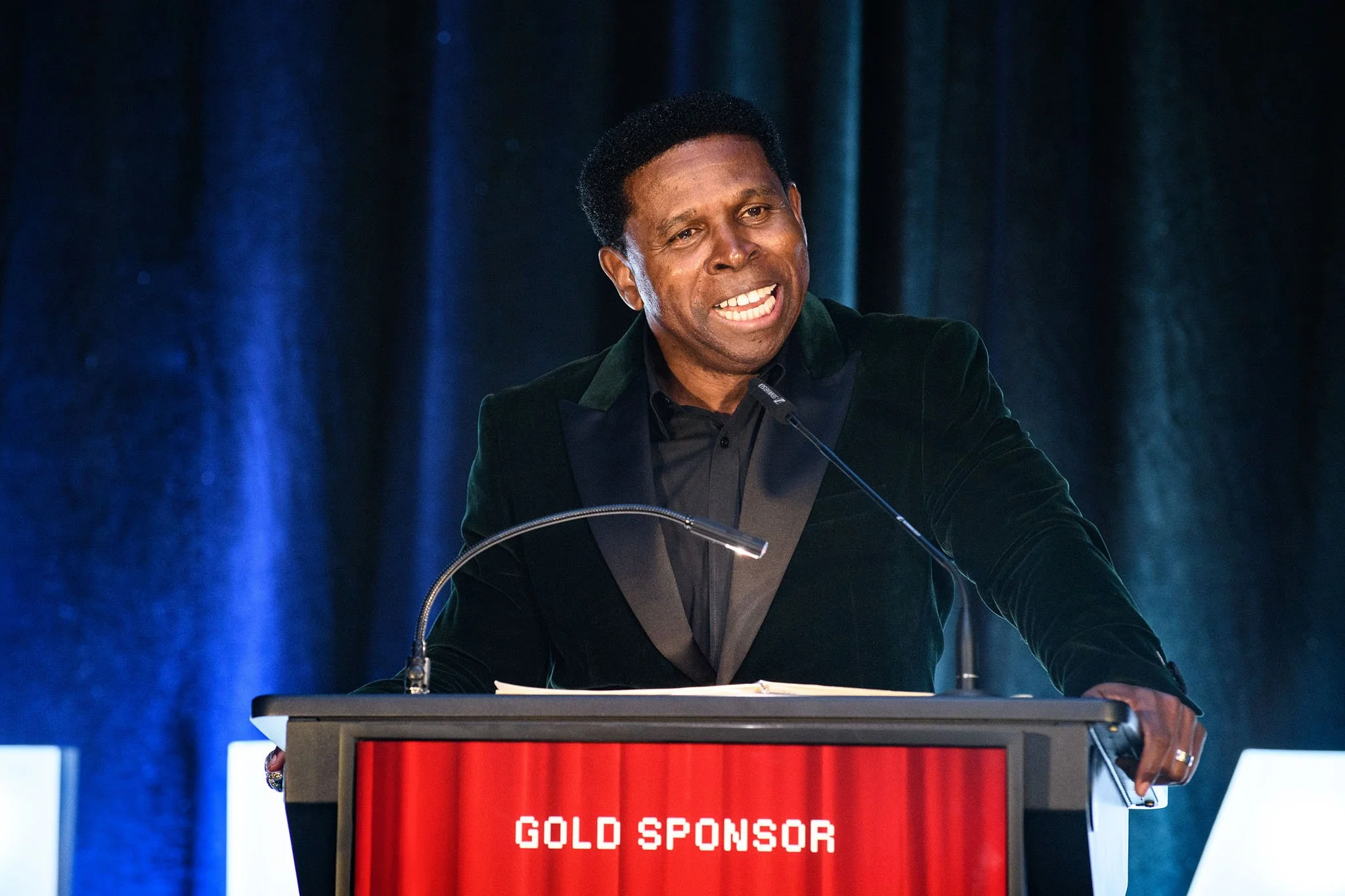 A man in a dark blazer and black shirt speaking at a podium during an event with a blue curtain in the background. The podium has a red digital display showing 'GOLD SPONSOR'.
