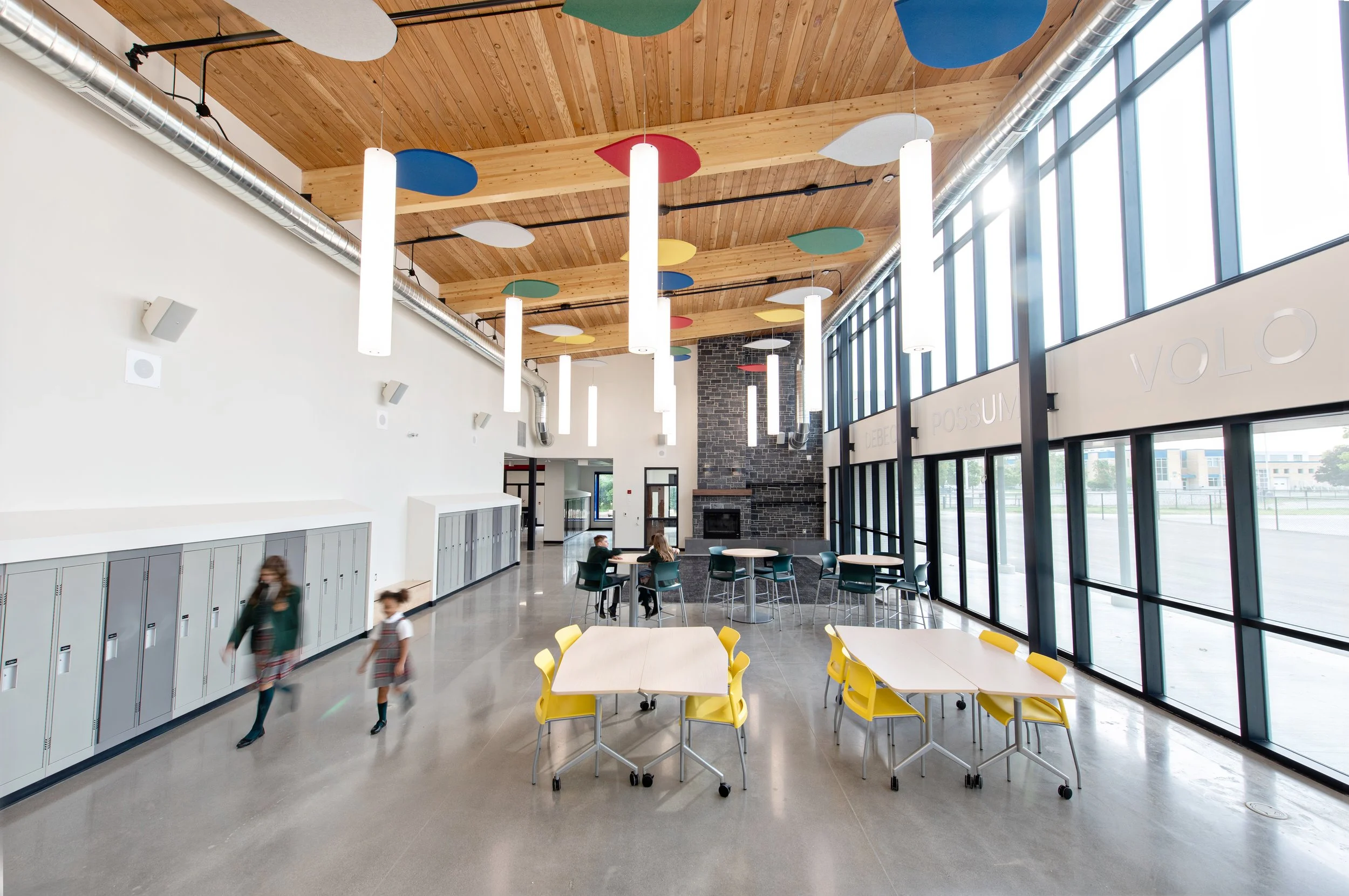 Interior of a modern school cafeteria with high ceiling, large windows, pendant lighting, and lockers along the wall. Children are walking and sitting at tables.