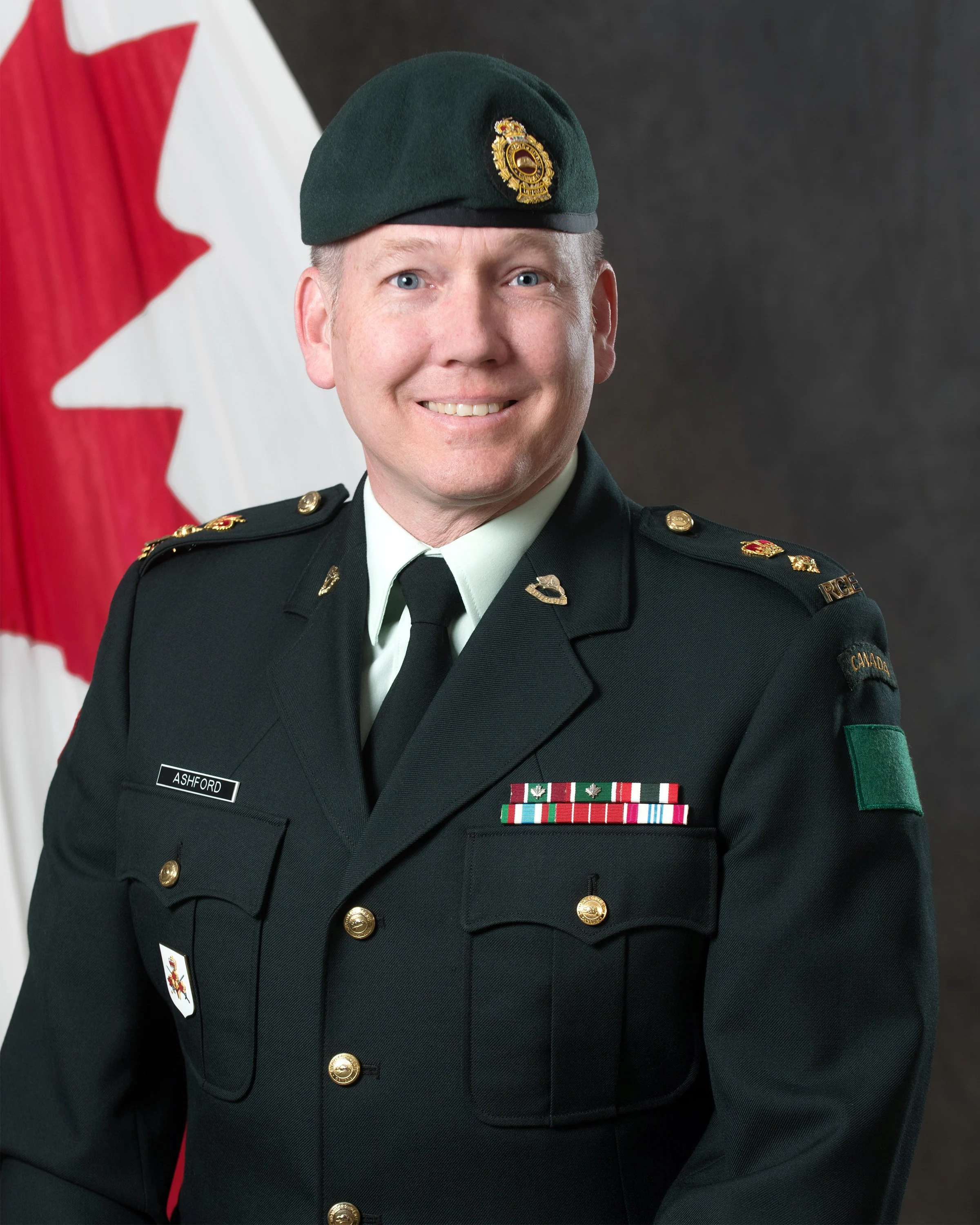 A uniformed officer in a black military uniform with medals and patches, wearing a dark green beret with a gold badge, standing in front of a Canadian flag.