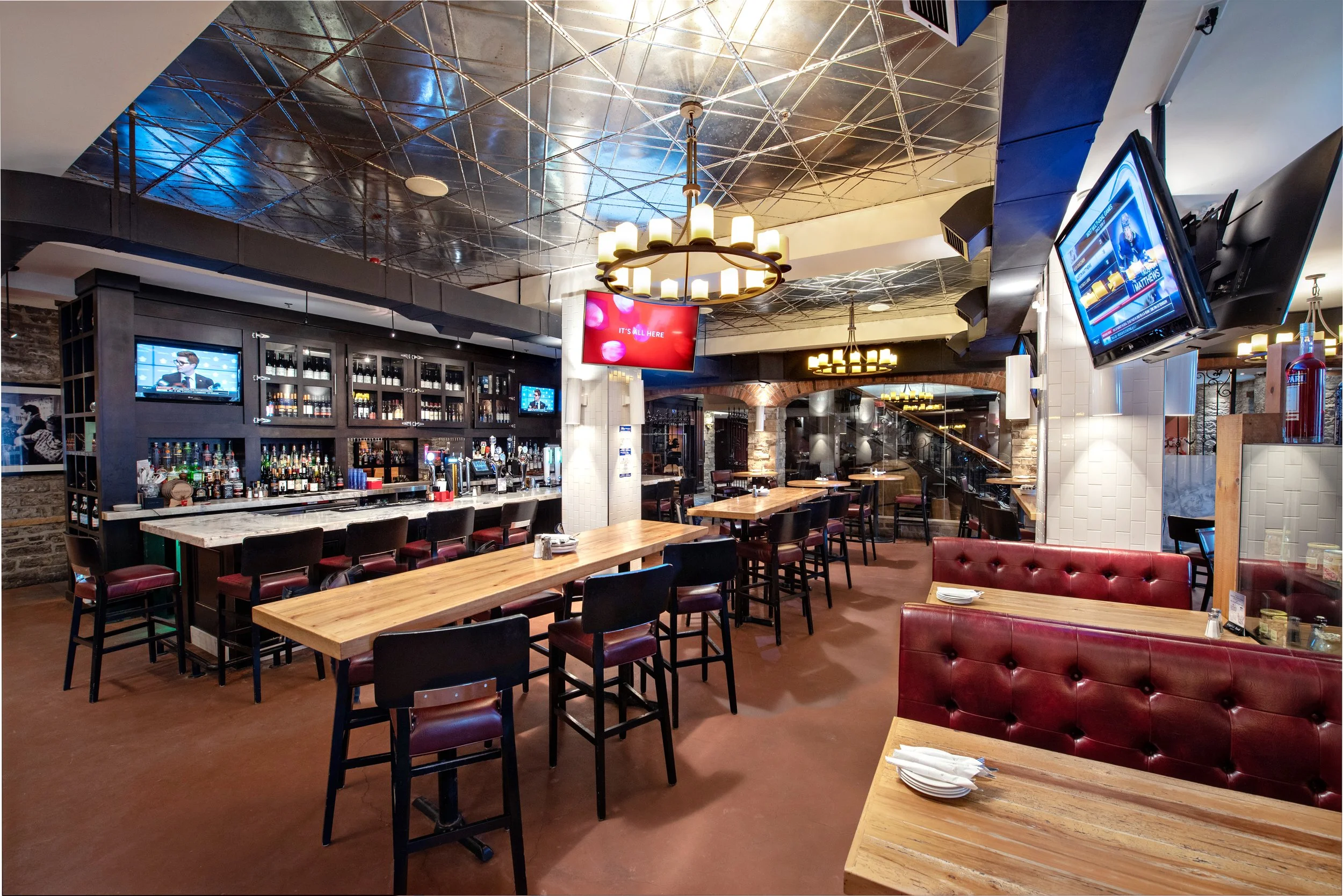 Interior of a restaurant or bar with wooden tables and red leather chairs, multiple wall-mounted televisions, a bar area with bottles, and modern decorative lighting fixtures. The ceiling has a reflective metallic surface.